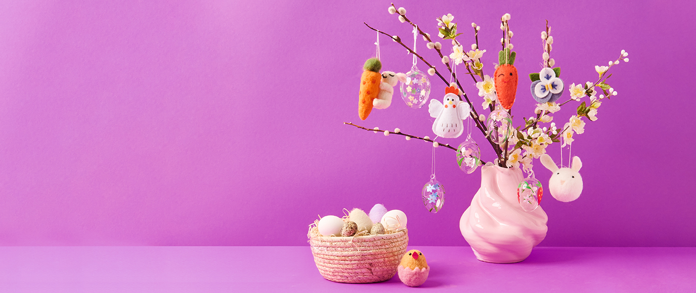 Weidenkätzchen in einer rosa Vase mit Filzfiguren und Glas-Ostereiern behangen, daneben ein Osterkörbchen mit verschiedenen dekorativen Ostereiern. 