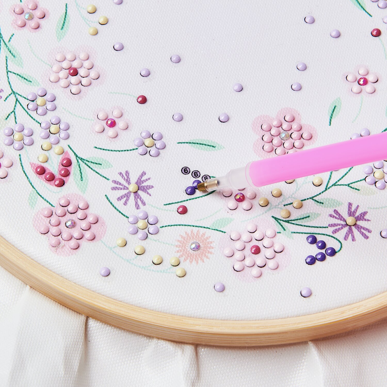 Diamond Painting DIY-Set Funkelnder Blumenkranz: Pastellfarbene Blumen auf weißem Stoff, Ø20cm, mit pinkem Stift verziert.