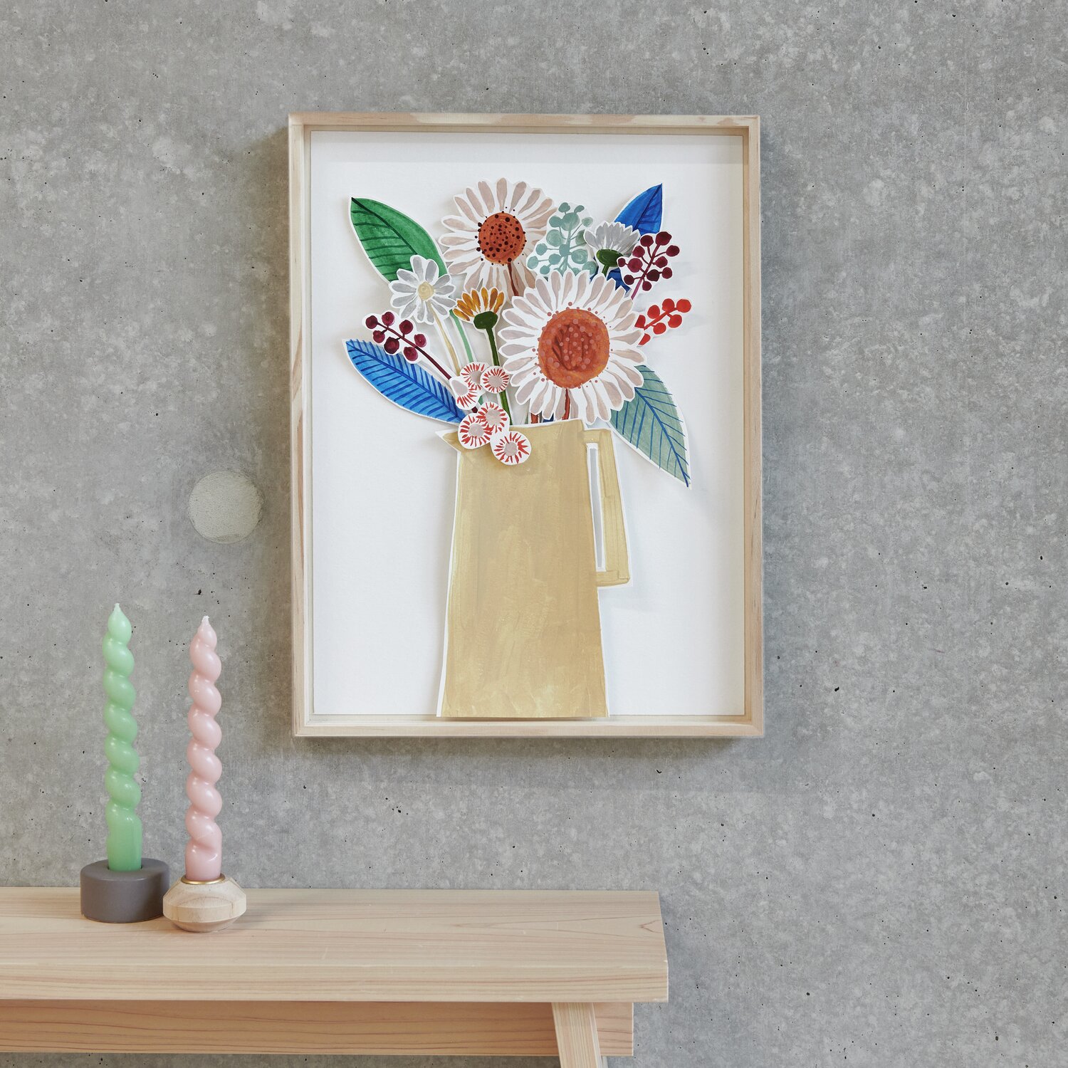 Blumenbild in Holzrahmen an grauer Wand, darunter pastellfarbene Kerzen auf Holzregal.