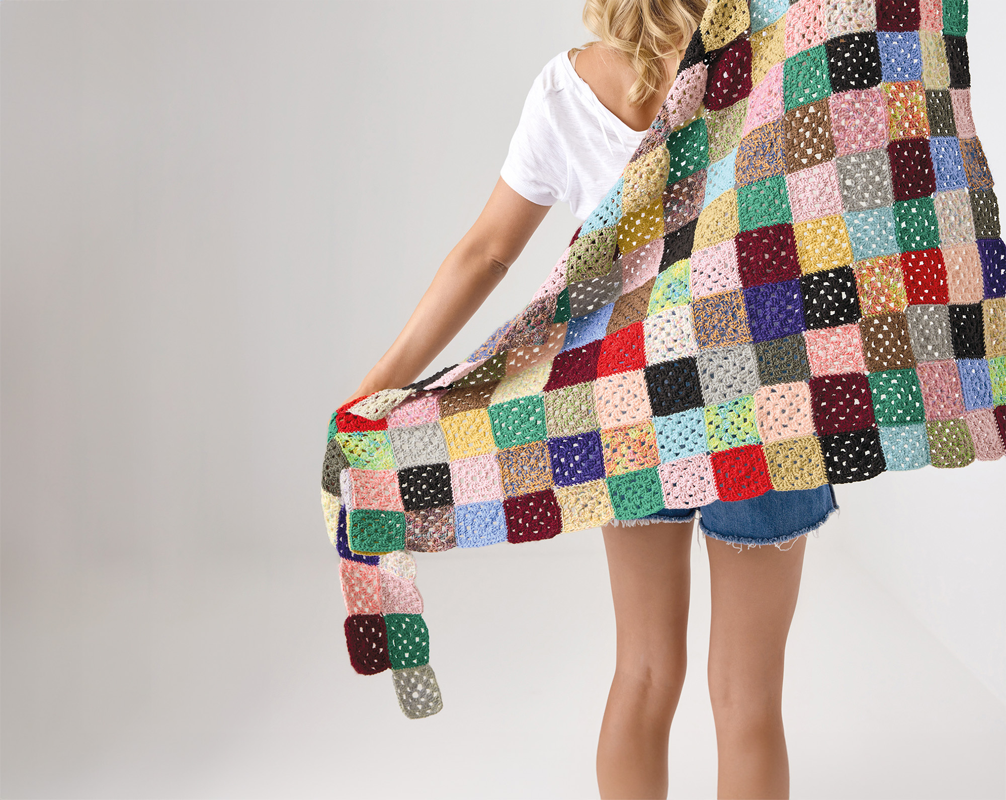 Eine, von hinten zusehende, Frau hält eine gehäkelte Decke aus bunten Granny-Squares hinter ihrem Rücken. Fotosetting weiße Wand.
