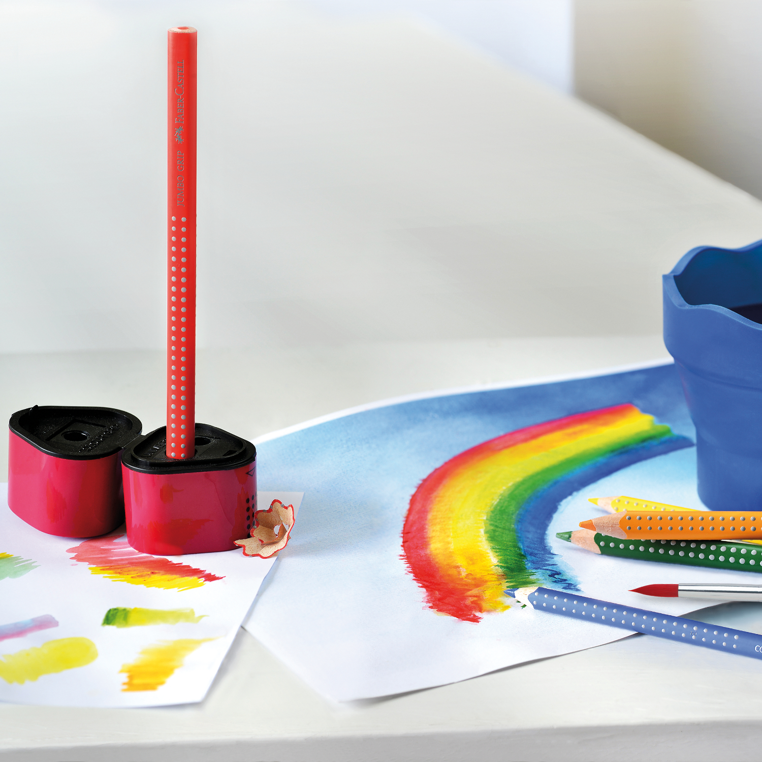 Jumbo Grip Farbstift in Purpurrosa von Faber Castell, stehend in einem Anspitzer, neben bunten Zeichnungen und weiteren Stiften.