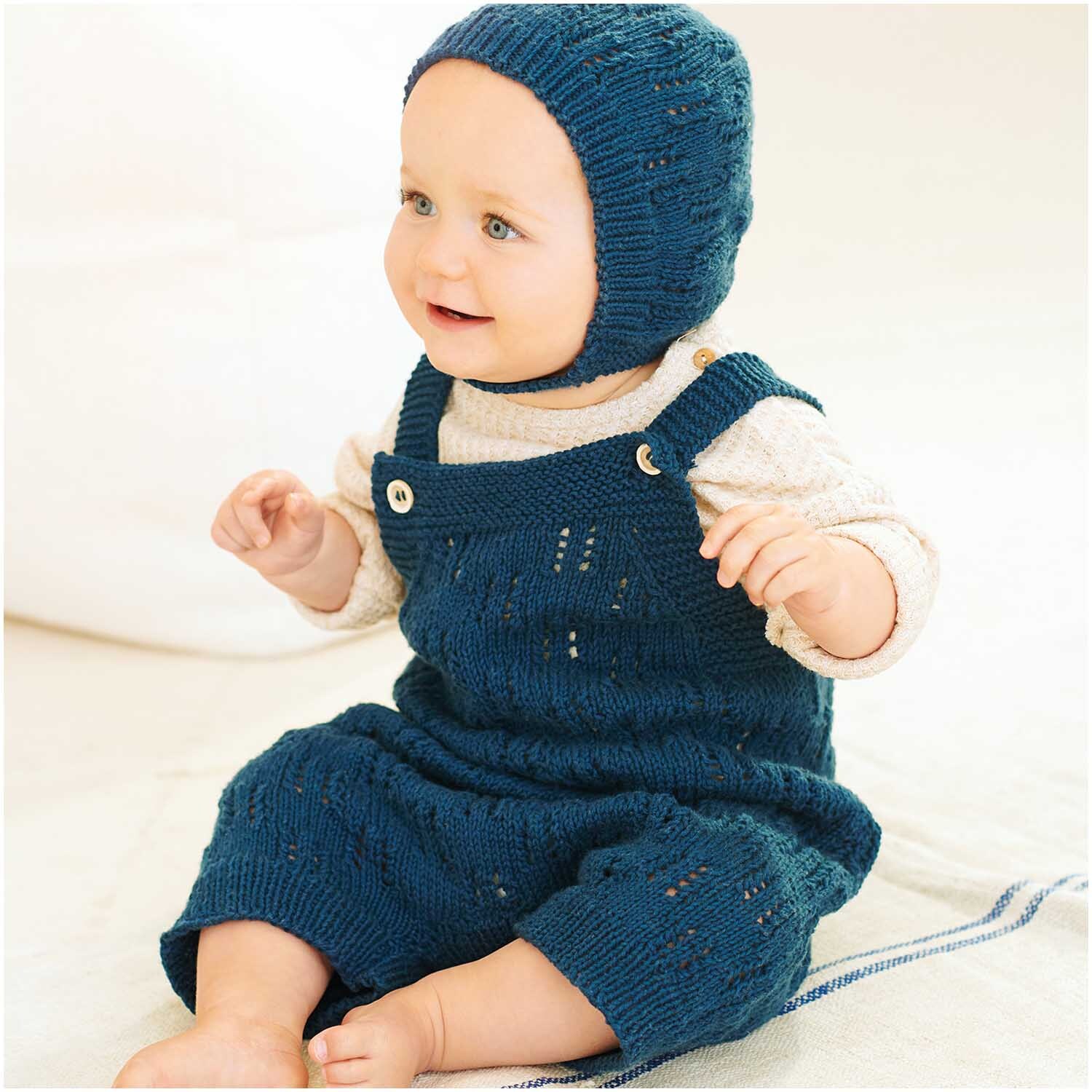 Baby Organic Cotton 50g 150m Farbe Marine von Rico Design Baby in gestricktem marineblauem Outfit aus Bio-Baumwolle von Rico Design, sitzt lächelnd auf hellem Hintergrund.