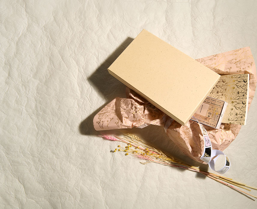 Geöffnete Geschenkbox mit dekorativem Seidenpapier, Papierprodukten und Trockenblumen auf hellem Hintergrund.