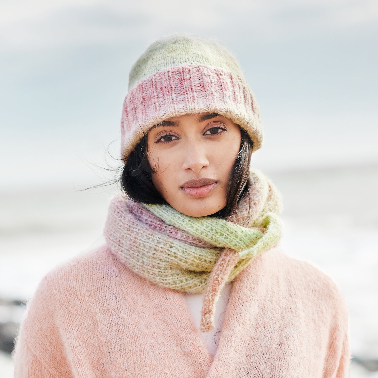 Strickset Tuch Modell 37 aus Lovewool No. 21 von Rico Design Strickset Mütze Modell 38 aus Lovewool No. 21: Frau trägt pastellfarbene Mütze und Schal am Strand.