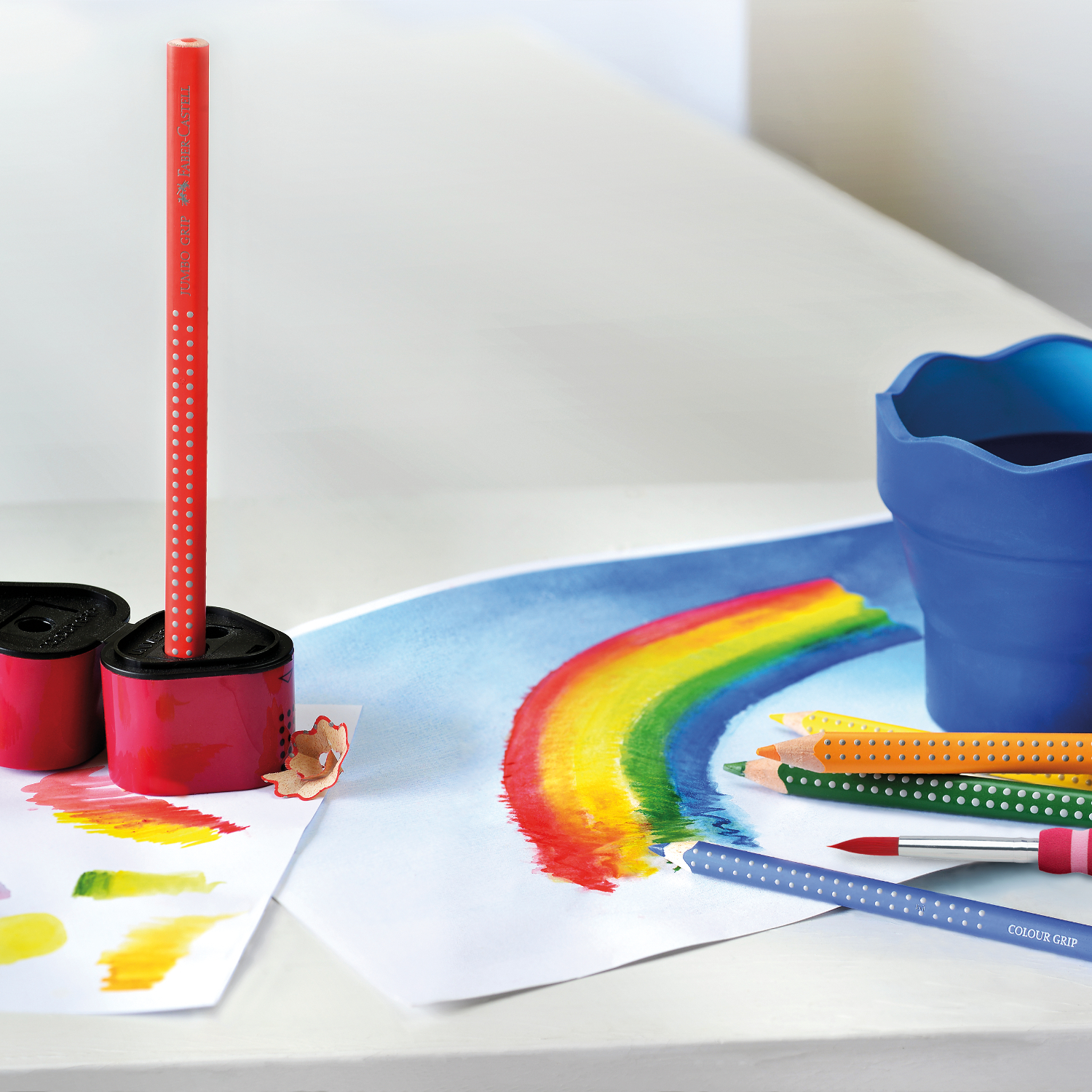 Jumbo Grip Farbstift, Faber Castell, auf Tisch mit Regenbogenzeichnung und blauem Becher.