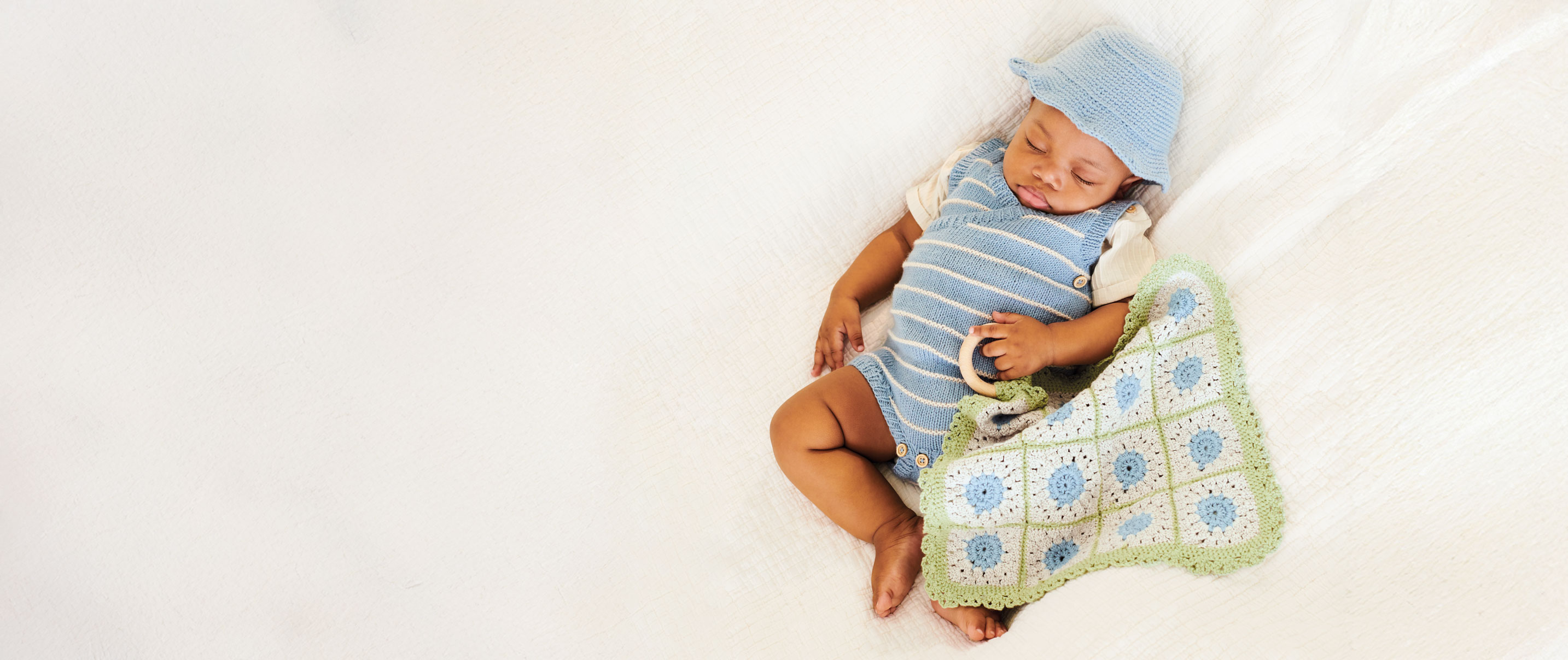 Baby liegt in Strickstrampler mit Häkeldecke auf Bett.