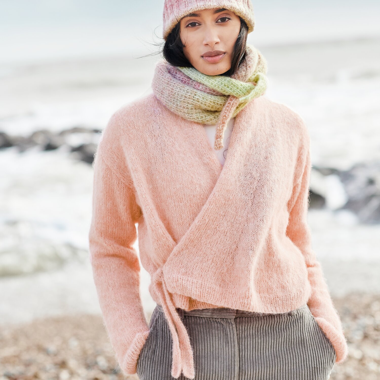 Strickset Jacke Langarm Modell 14b aus Lovewool No. 21 von Rico Design Strickset Jacke Langarm Modell 14b aus Lovewool No. 21: Frau am Strand in rosa Strickjacke, kombiniert mit Schal und Mütze in Pastelltönen.