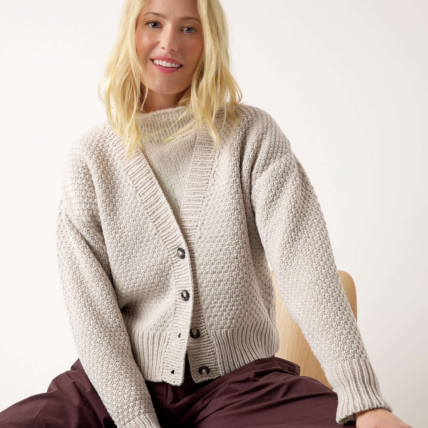 Strickset Cardigan Modell 01 aus Die Neue Masche No. 13 von idee. Creativmarkt Strickset Cardigan Modell 01: Frau trägt beigen Cardigan mit Knöpfen, sitzt entspannt auf Stuhl.