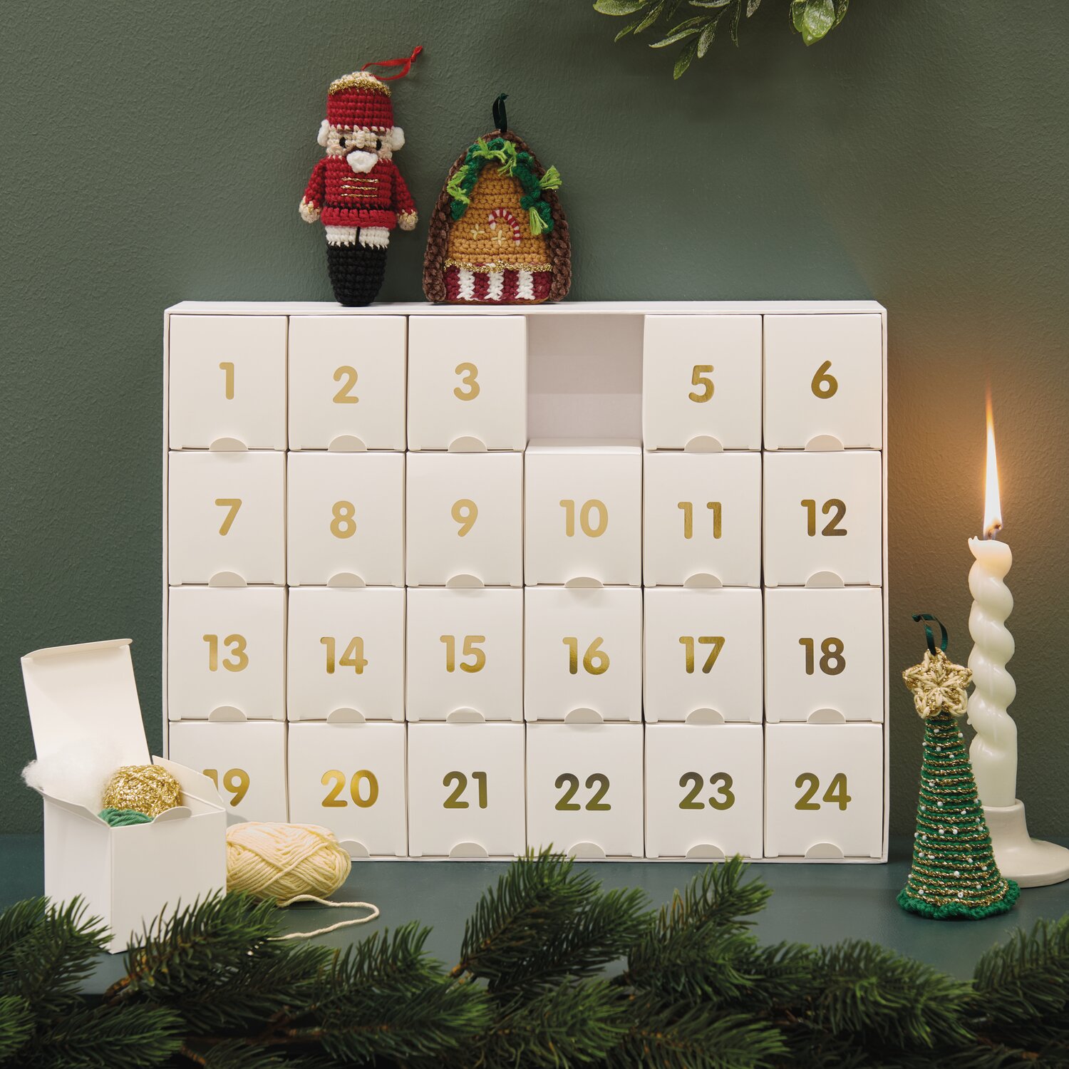 Ricorumi Adventskalender Weihnachtsmarkt mit weißen Boxen und goldenen Zahlen, dekoriert mit gehäkelten Figuren und Kerzen auf grünem Hintergrund.