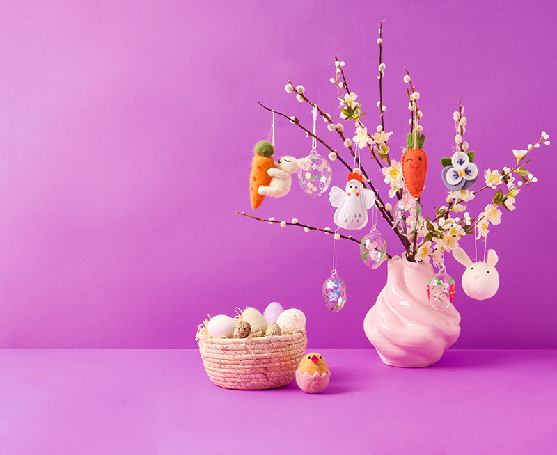 Weidenkätzchen in einer rosa Vase mit Filzfiguren und Glas-Ostereiern behangen, daneben ein Osterkörbchen mit verschiedenen dekorativen Ostereiern. 
