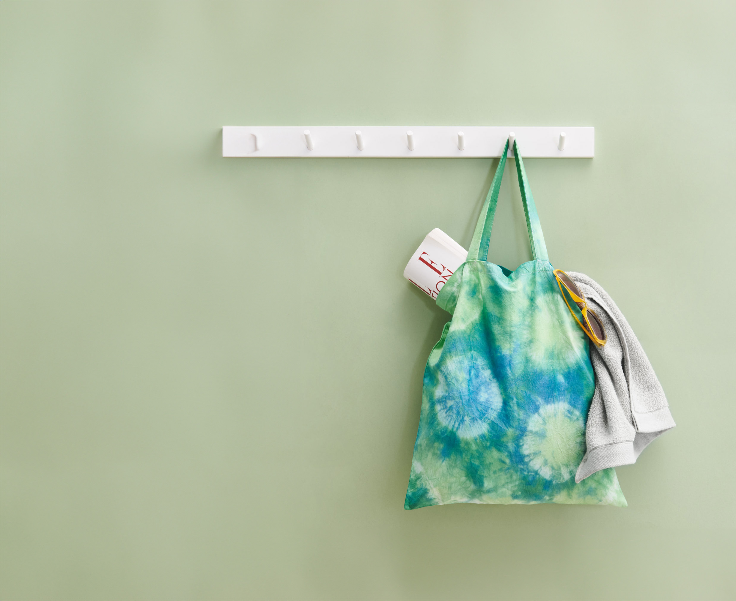 Ein Beutel mit blau-grünem Batik-Design hängt an einer weißen Garderobe. Fotosetting grüne Wand.