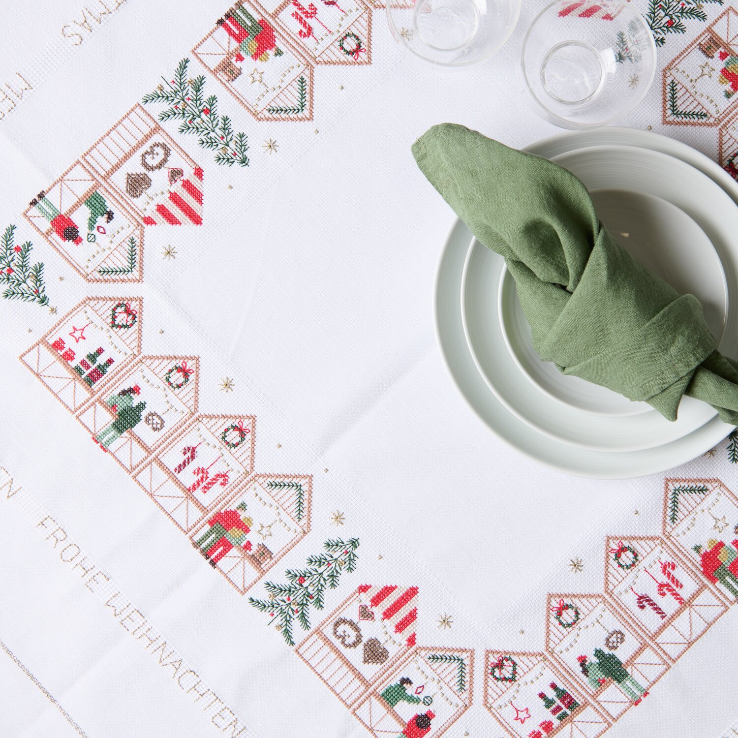 Stickpackung Tischdecke Weihnachtsmarkt: Festliche Tischdecke mit Weihnachtsmotiven, 90x90cm, von Rico Design, dekoriert mit Geschirr und grünem Stoffserviette.