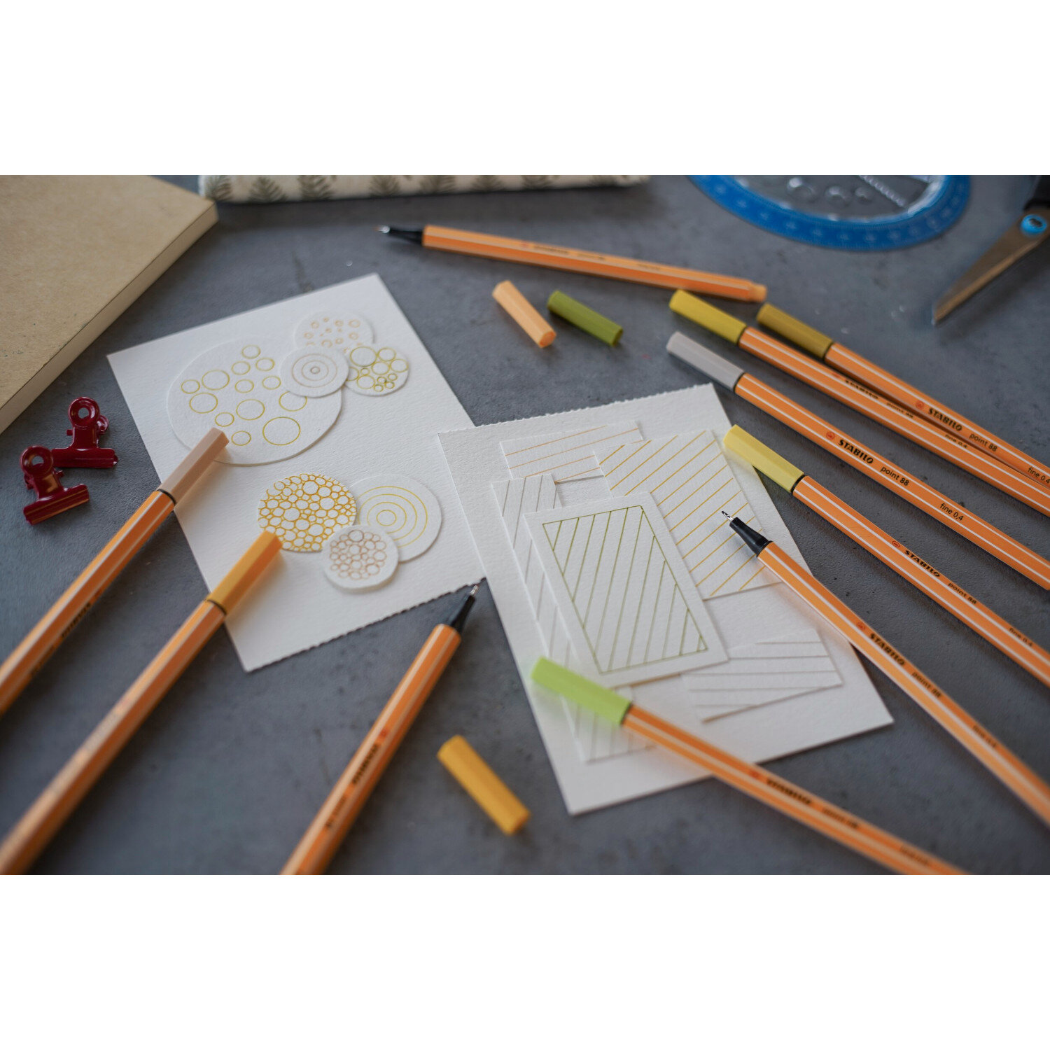 Point 88 Fineliner Pastell im Etui 8 Farben auf einem Tisch mit Zeichnungen und Notizbuch.