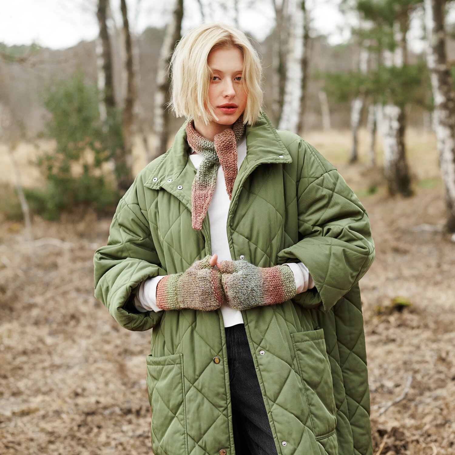 Strickset Minituch Modell 04 aus Lovewool No. 21 von Rico Design Strickset Minituch Modell 04 aus Lovewool No. 21: Frau im grünen Mantel mit gestricktem Schal und Handschuhen in Naturtönen im Wald.