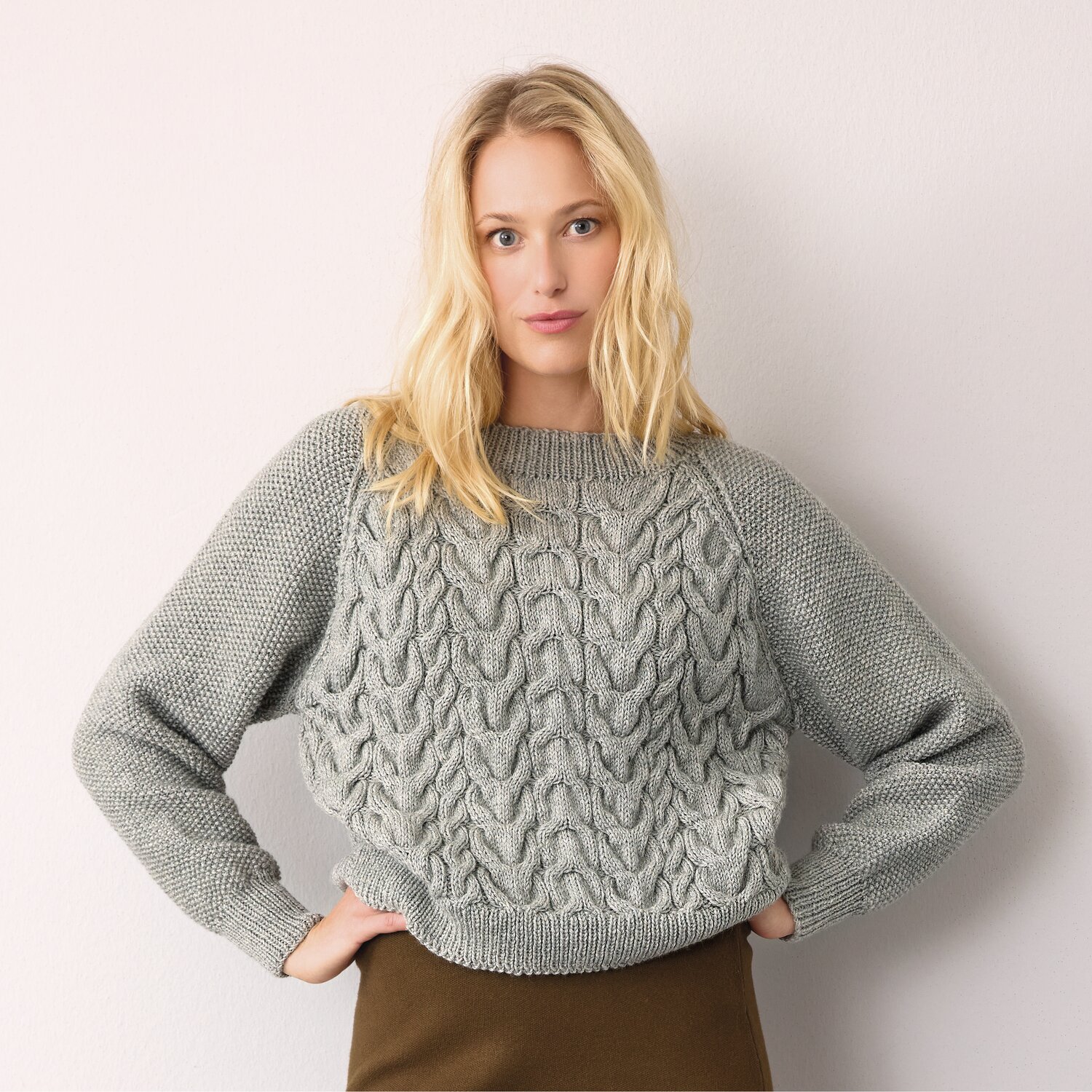 Strickset Pullover Modell 27 aus Die Neue Masche No. 14 von idee. Creativmarkt Strickset Pullover Modell 27 aus DNM 14: Grauer Pullover mit Zopfmuster, getragen von Frau vor hellem Hintergrund.