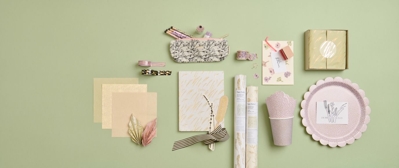 Flach ausgelegte Papeterie- und Geschenkartikel in Pastellfarben auf hellgrünem Hintergrund, darunter Notizbuch, Geschenkverpackungen, Karten, Geschenkpapier und dekorative Accessoires.