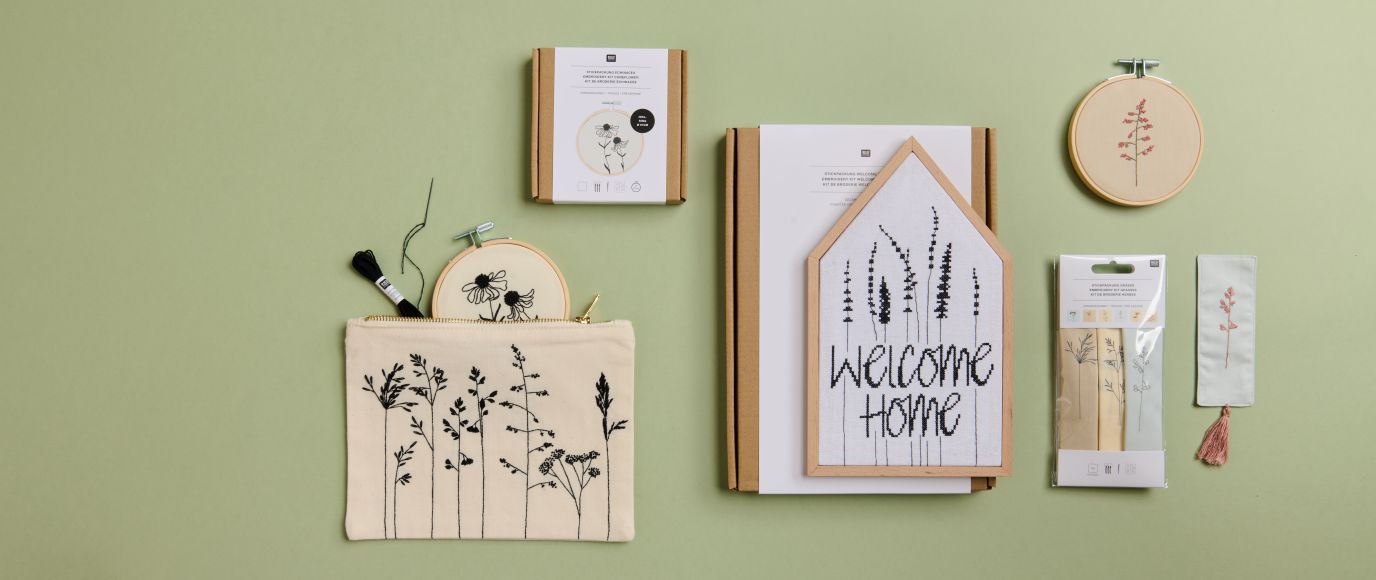 Naturfarbene Stofftasche, Reißverschlusstasche und Stickrahmen mit floralen Motiven vor einem hellgrünen Hintergrund.