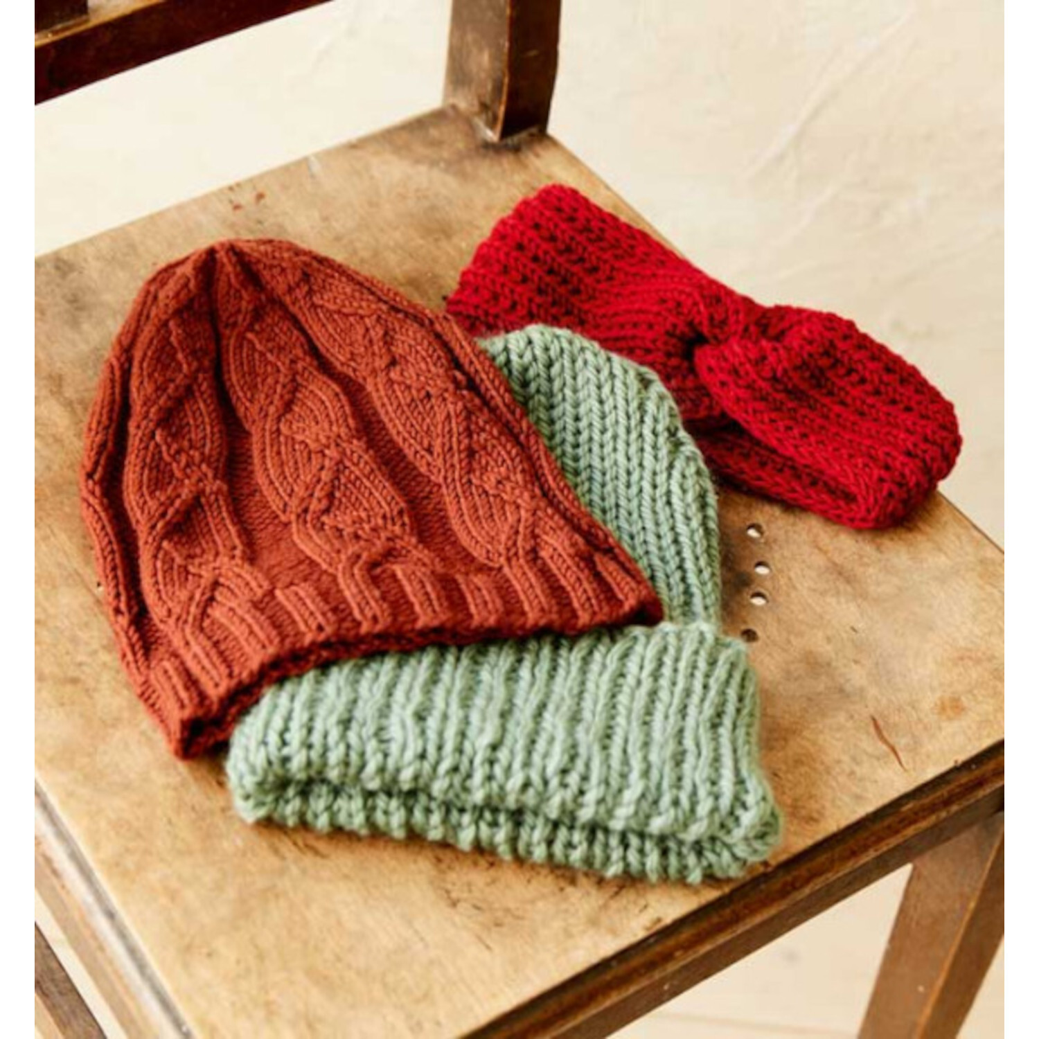 Stricken mit Struktur: Drei gestrickte Mützen in Rot, Braun und Grün auf einem Holzstuhl. Ideal für kalte Tage.