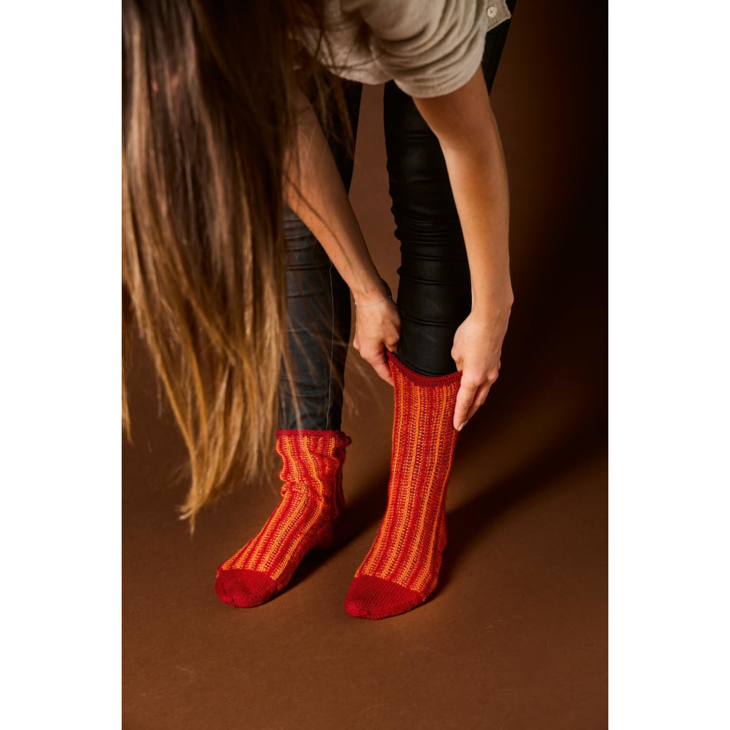 Socken stricken mit Wow-Effekt: Frau zieht rote, gestreifte Socken an, kniend auf braunem Boden.