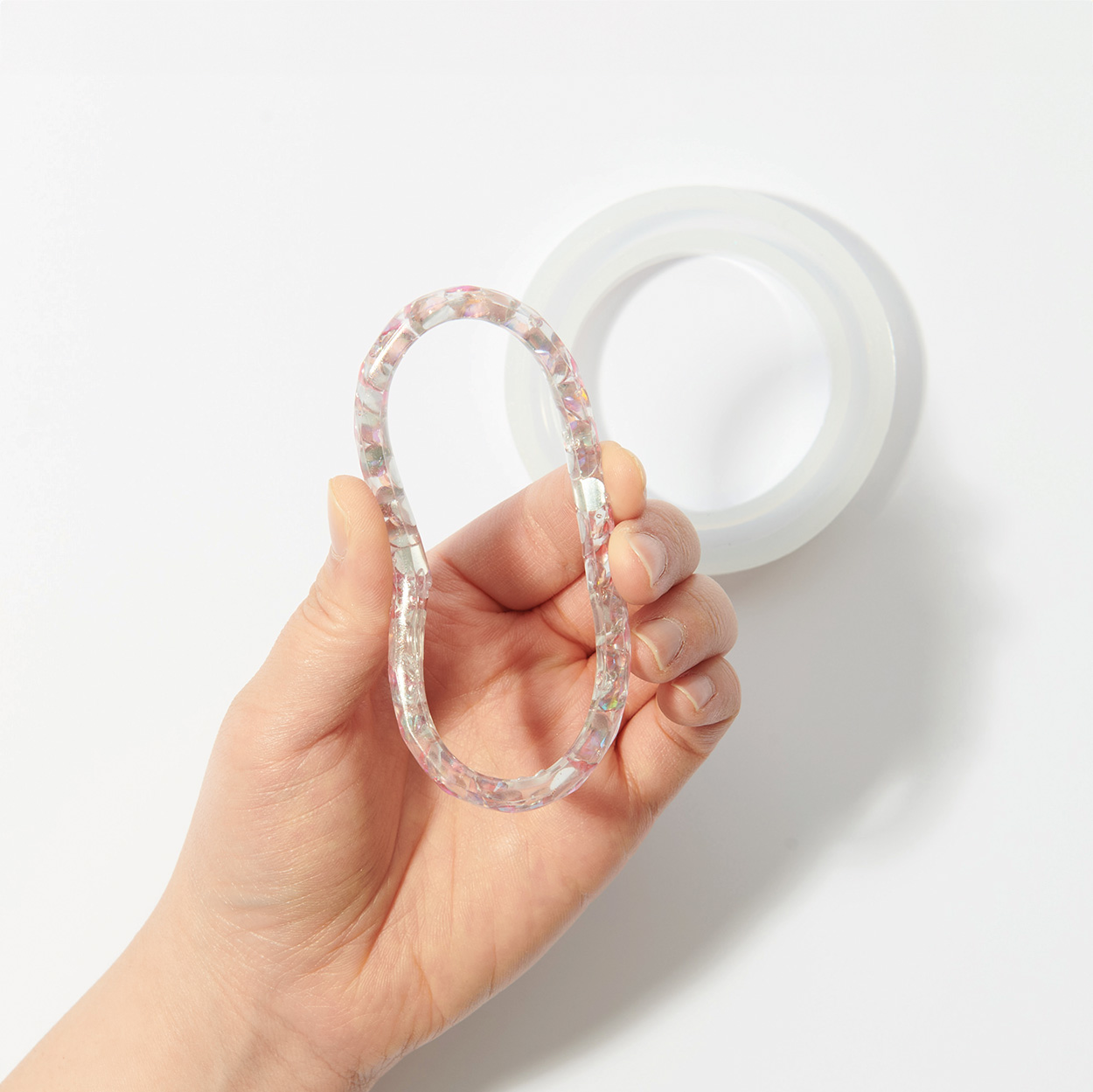 Hand biegt einen transparenten Armreif mit Glitzer, darunter eine runde Silikonform. Flatlay von Produkten.