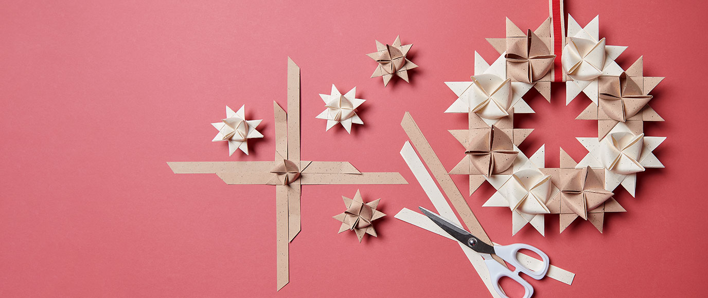 Ein Kranz aus hell- und dunkelbeigen Fröbelsternen, daneben eine Schere, Papierstreifen und weitere Fröbelsterne. Flatlay von Produkten.