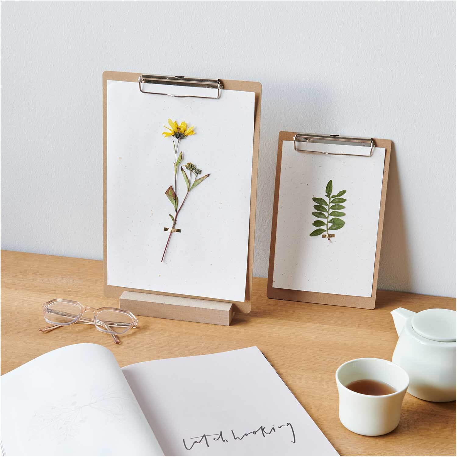 Klemmbrett Natur DIN A4 mit gepresster Blume, auf Holzunterlage, neben Brille und Teetasse.