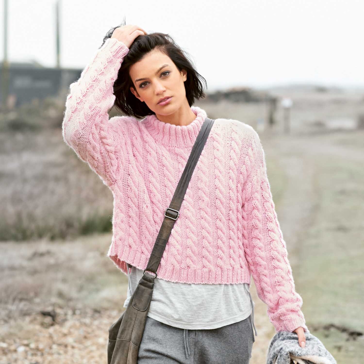 Strickanleitung Pullover aus Essentials Alpaca Blend chunky: Frau trägt rosa Zopfpullover in ländlicher Umgebung.