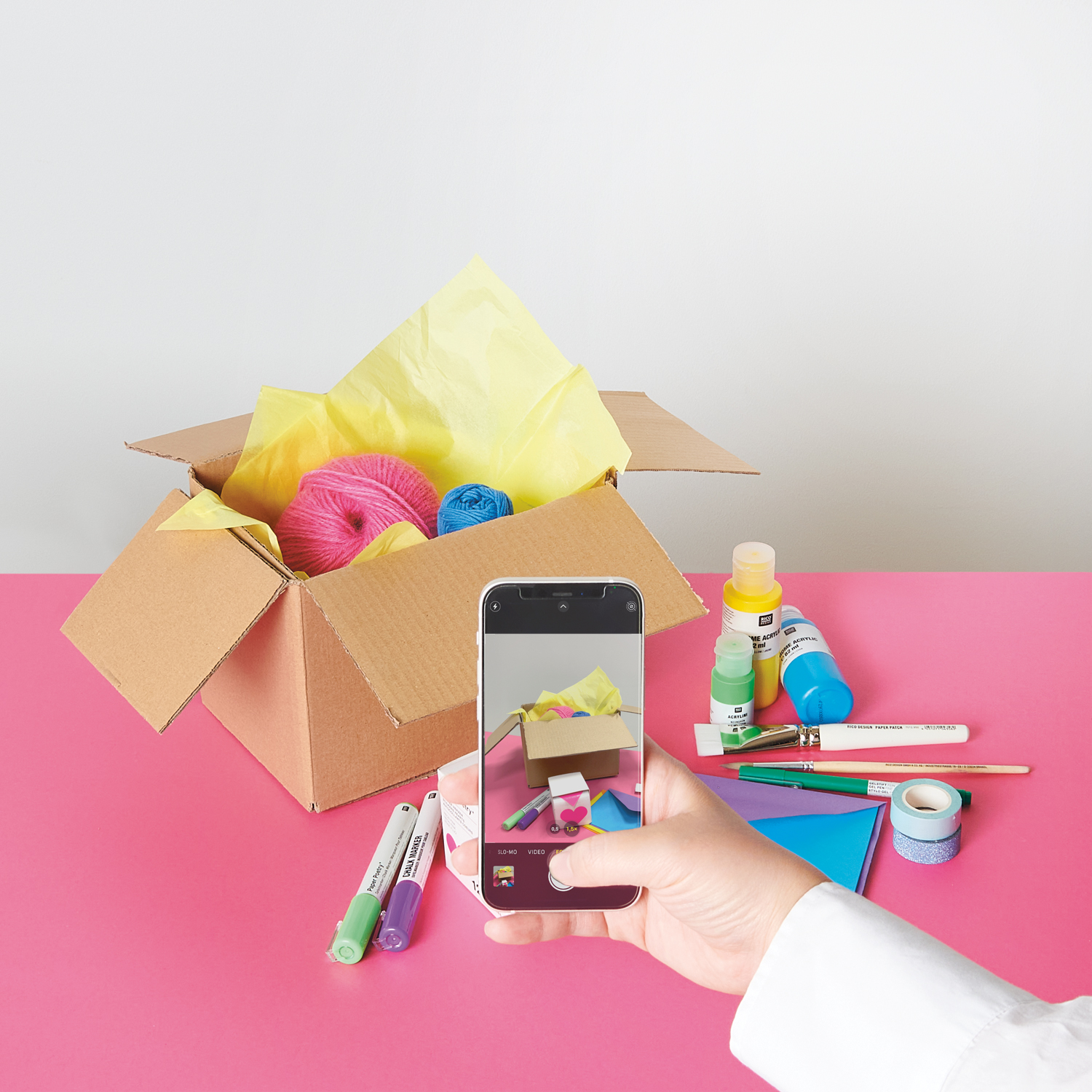 Ein Karton mit Wolle drin, daneben Stifte, Farben, Pinsel und Tapes, im Vordergrund eine Hand mit Handy, welche ein Foto von den Produkten macht.