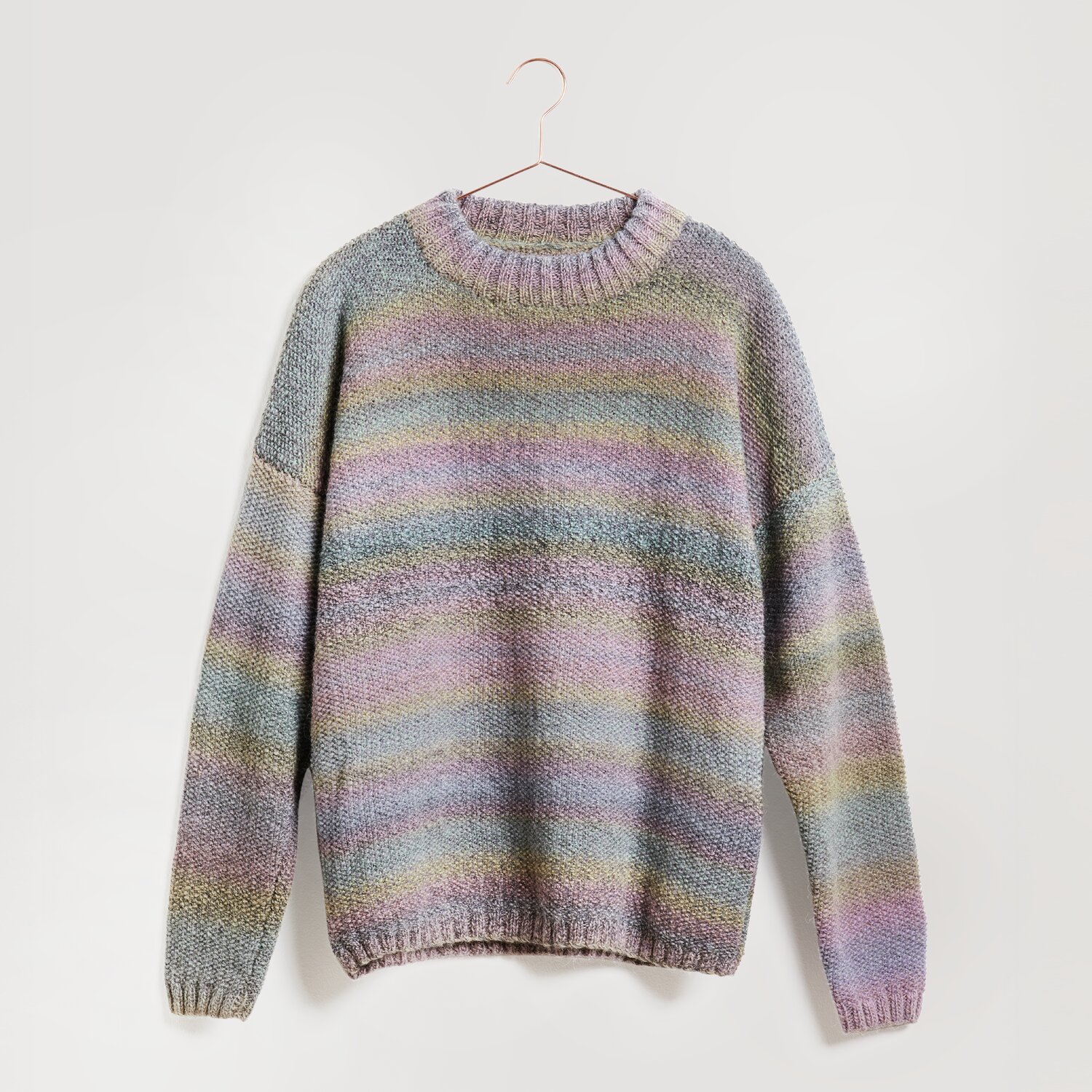 Lovewool No. 21 Herbst-Winter Sprachauswahl deutsch von Rico Design Ein gestrickter Pullover in sanften Pastelltönen hängt an einem Kleiderbügel. Der Pullover zeigt ein Streifenmuster in Blau, Rosa und Grün. Marke: Modell Legeware. Perfekt für einen gemütlichen Look.