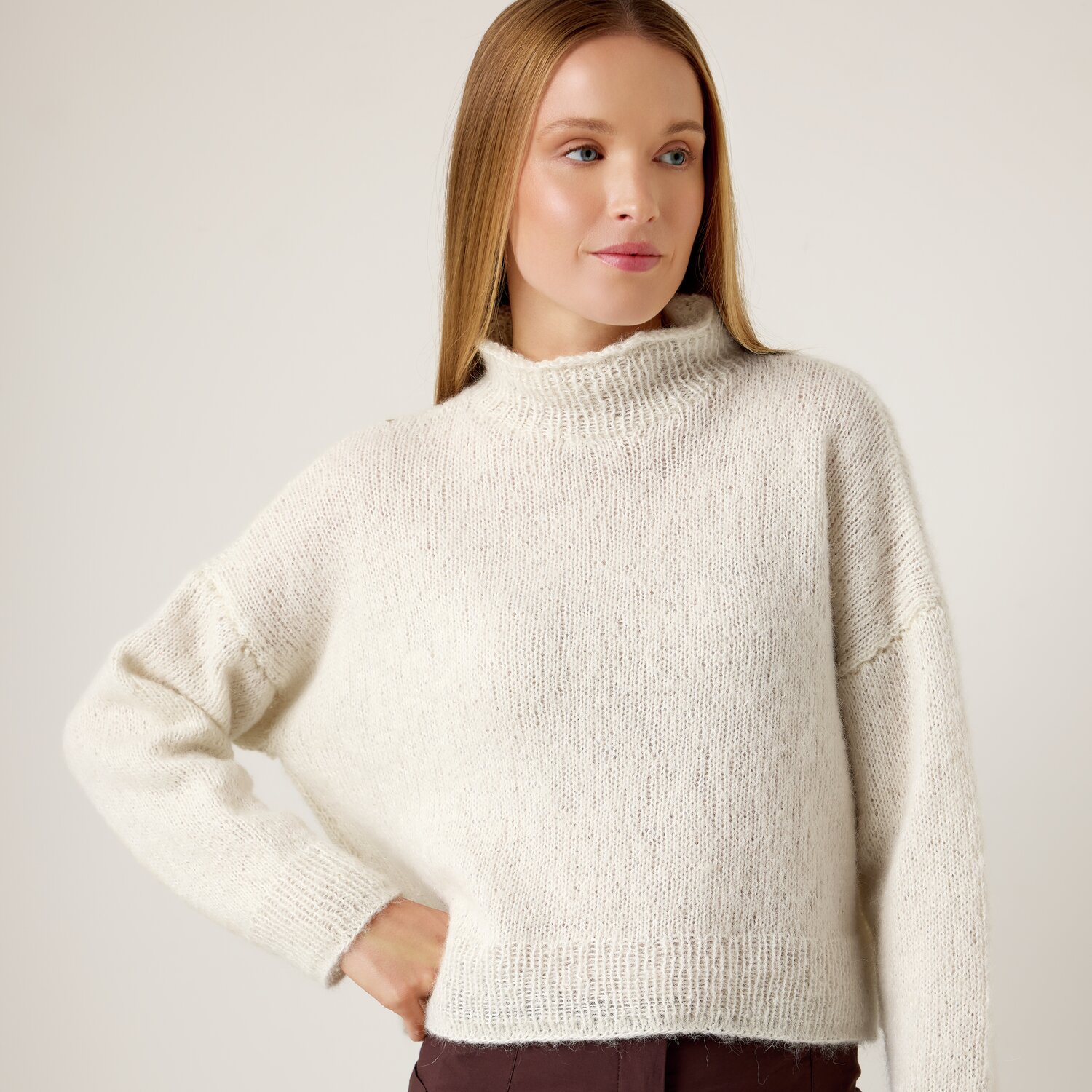 Strickset Pullover Modell 02 aus Die Neue Masche No. 13 von idee. Creativmarkt Strickset Pullover Modell 02 aus Die Neue Masche No. 13, cremefarbener Pullover mit hohem Kragen, getragen von einer Frau vor neutralem Hintergrund.