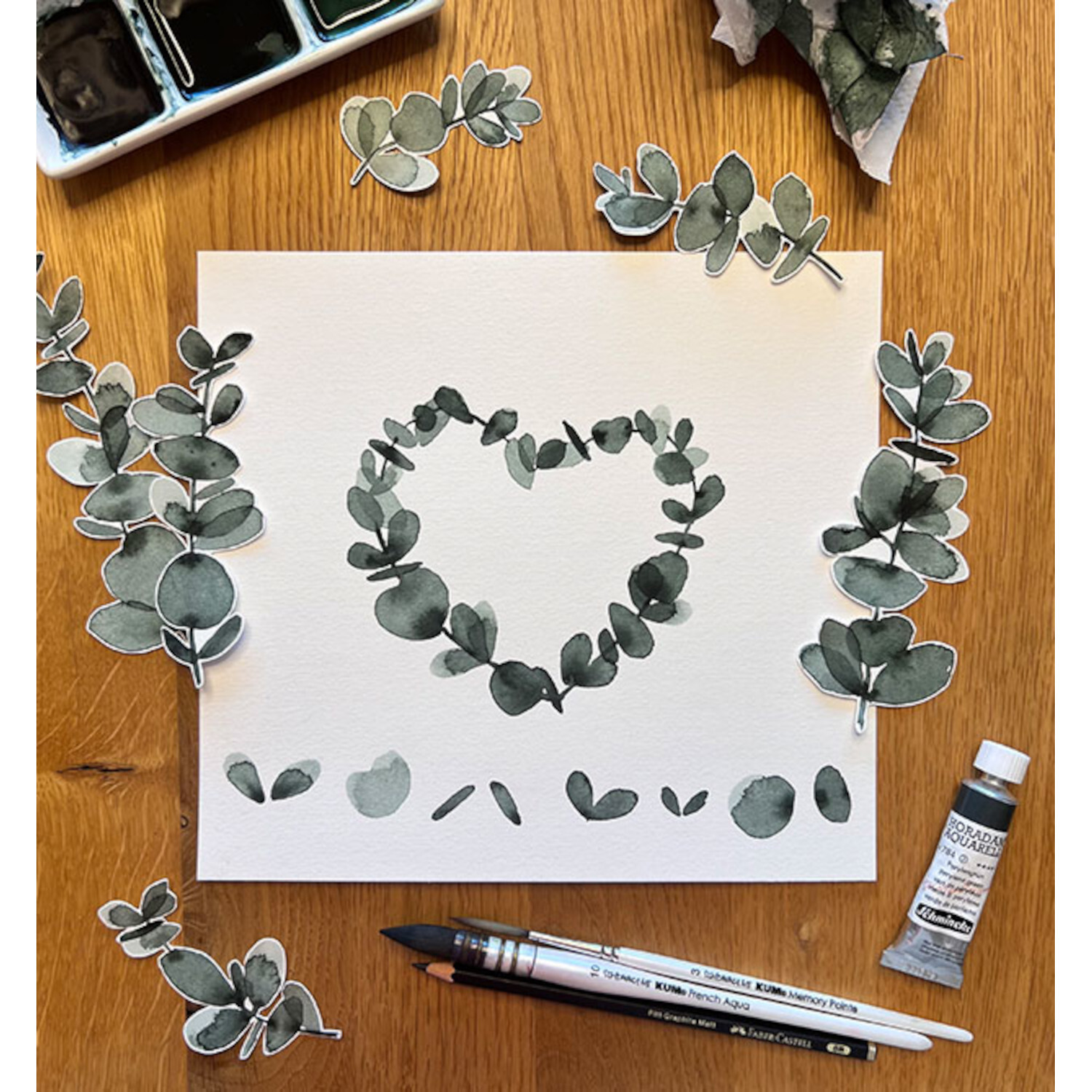 Watercolor your Mind: Herz aus grünen Blättern auf Papier, umgeben von Malutensilien auf Holztisch.