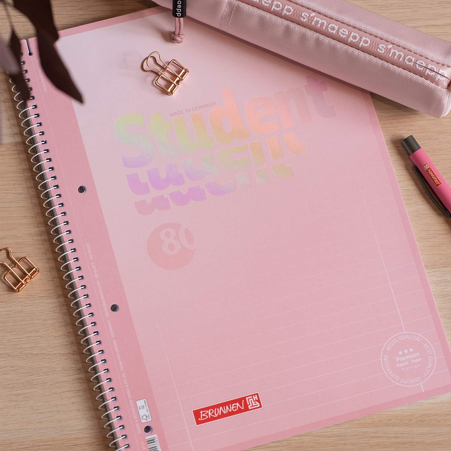 Collegeblock Pastell Rosa liniert 90g zeigt auf einem Schreibtisch einen Spiralblock von Brunnen mit 80 Blatt A4 neben Mäppchen, Stiften und Büroklammern