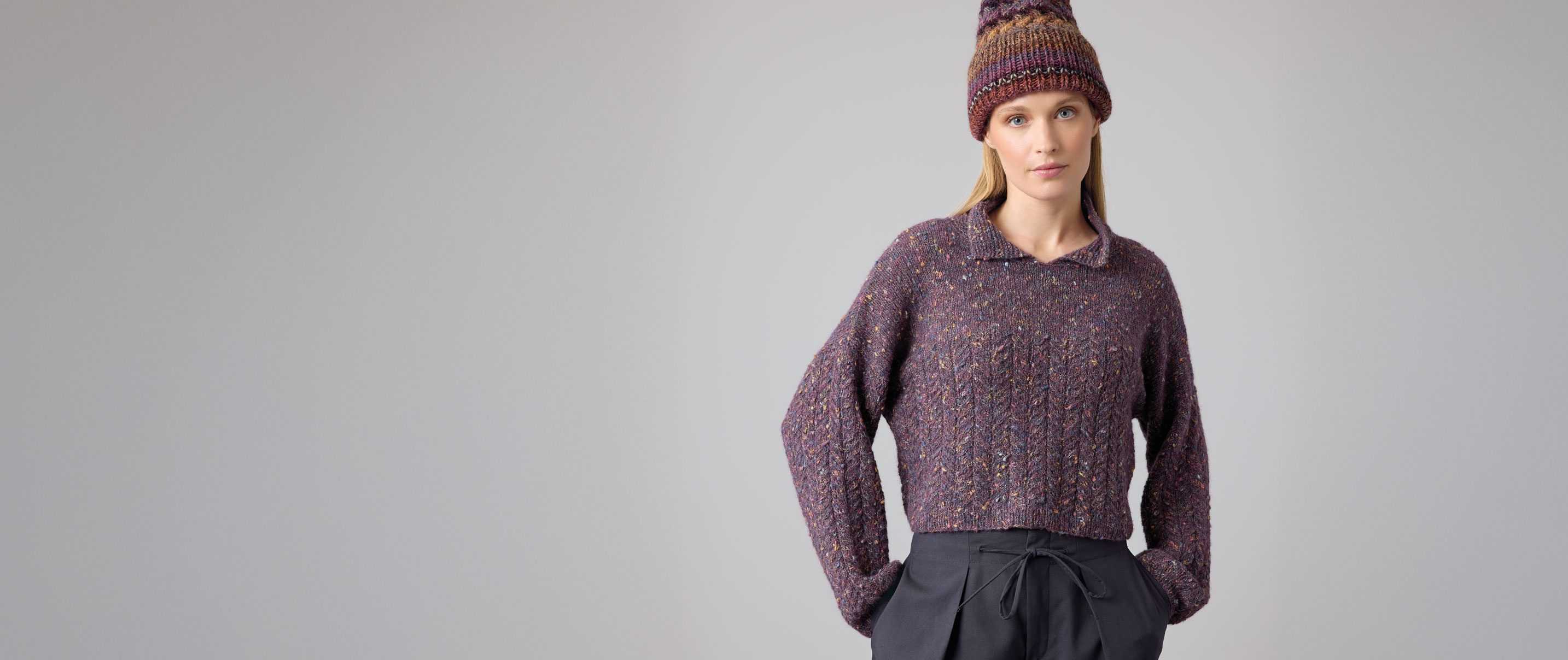 Frau trägt lilafarbenen Strickpulli und lilafarbene Strickmütze vor hellem Hintergrund