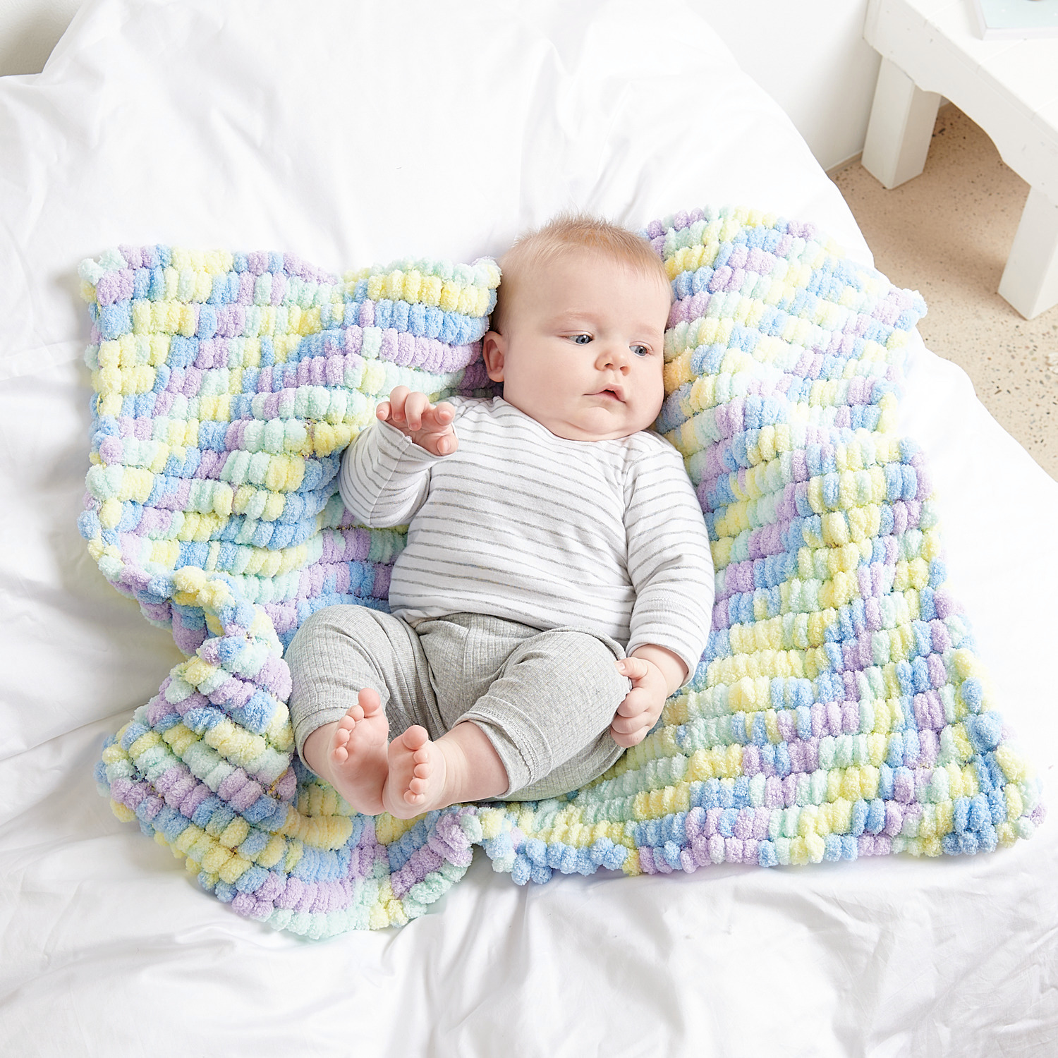 Creative Pompon Print: Ein Baby liegt auf einer weichen, bunten Decke von Rico Design in Blau Bunt.