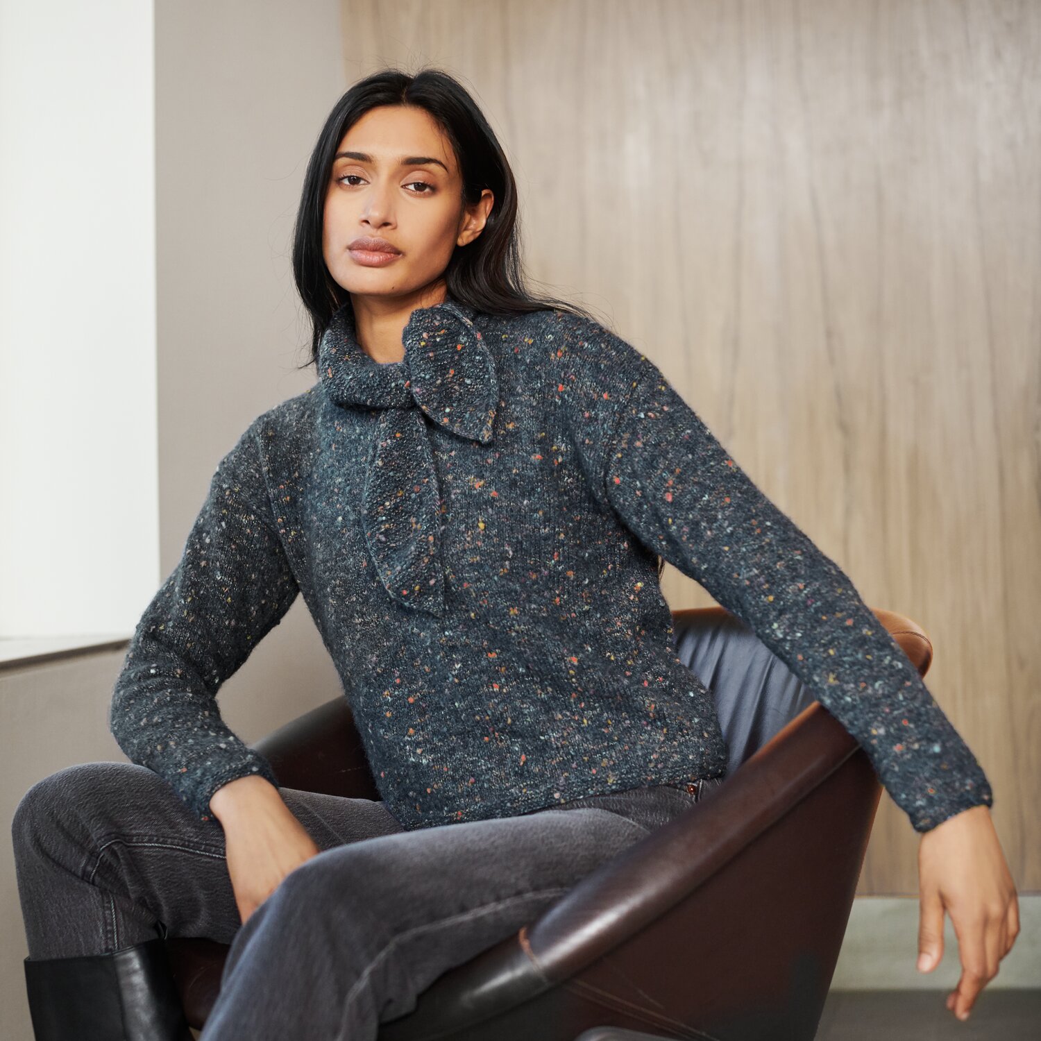 Strickset Pullover Modell 27 aus Lovewool No. 21 von Rico Design Strickset Pullover Modell 27 aus Lovewool No. 21, grauer Pullover mit Schleife, getragen von Frau auf Sessel.