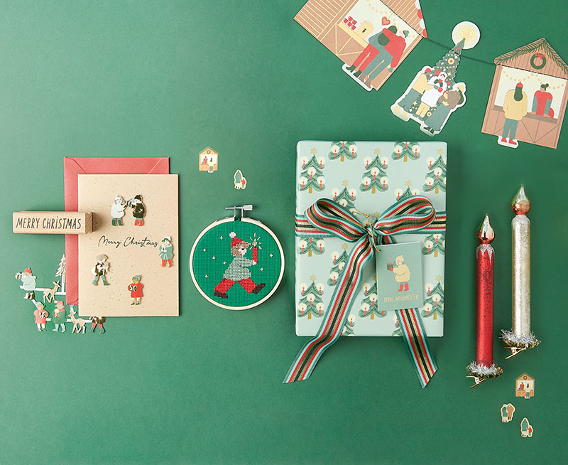 Ein verpacktes Geschenk, Geschenkpapierrollen, eine verzierte Weihnachtskarte, ein Stickring, eine verzierte Geschenkschachtel, Kerzen und eine Girlande alle in weihnachtlichem Stil. 