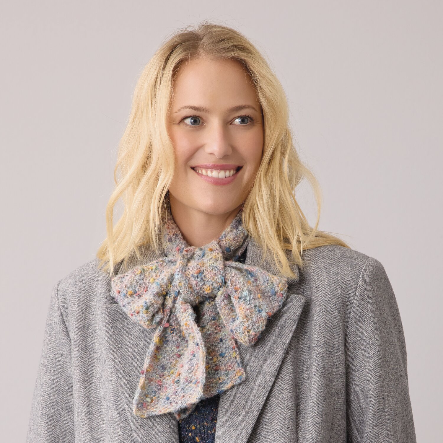 Strickset Mini-Schal Modell 14a aus Die Neue Masche No. 14 von idee. Creativmarkt Strickset Mini-Schal Modell 14a aus DNM 14: Frau trägt bunten Schal zu grauem Mantel. Ideal für stilvolle Herbstmode.