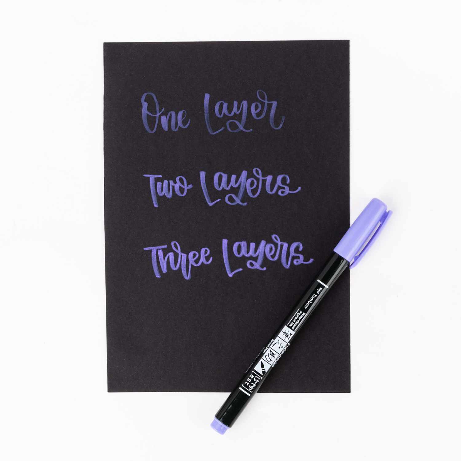 Fudenosuke Brush Pen Pastel von Tombow in Lavendel, neben schwarzem Papier mit Schriftzügen in verschiedenen Schichten.