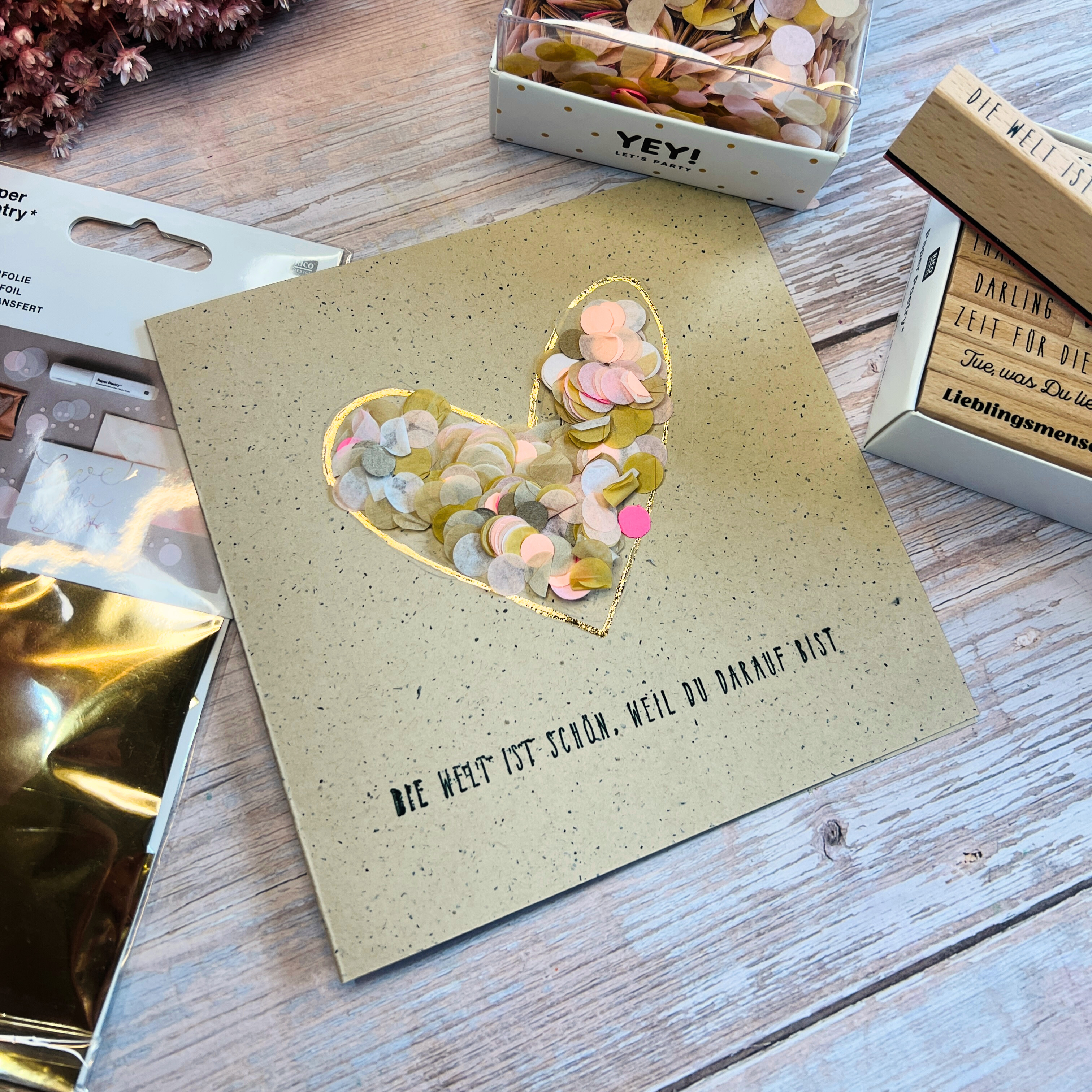 Eine quadratische Karte mit gold-umrandeten Konfetti-Herz und Schriftzug darunter.