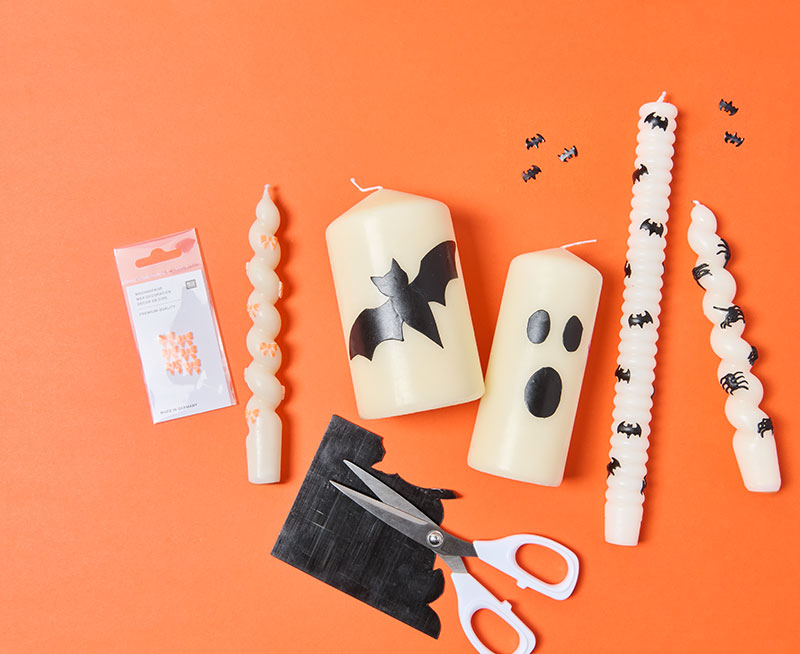 selbstgestaltete Kerzen mit Halloweenmotiven vor einem orangefarbenen Hintergrund.