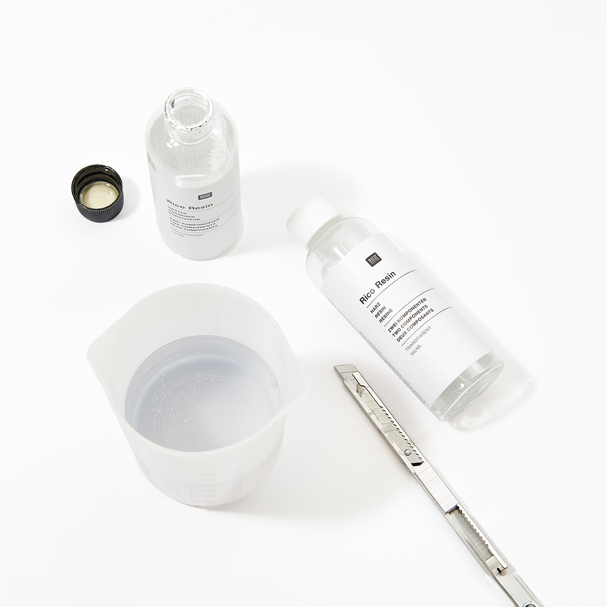 Epoxidharz in einem durchsichtigen Messbecher, daneben eine stehende und eine liegende Flasche Epoxidharz mit einem Cuttermesser. Flatlay von Produkten.