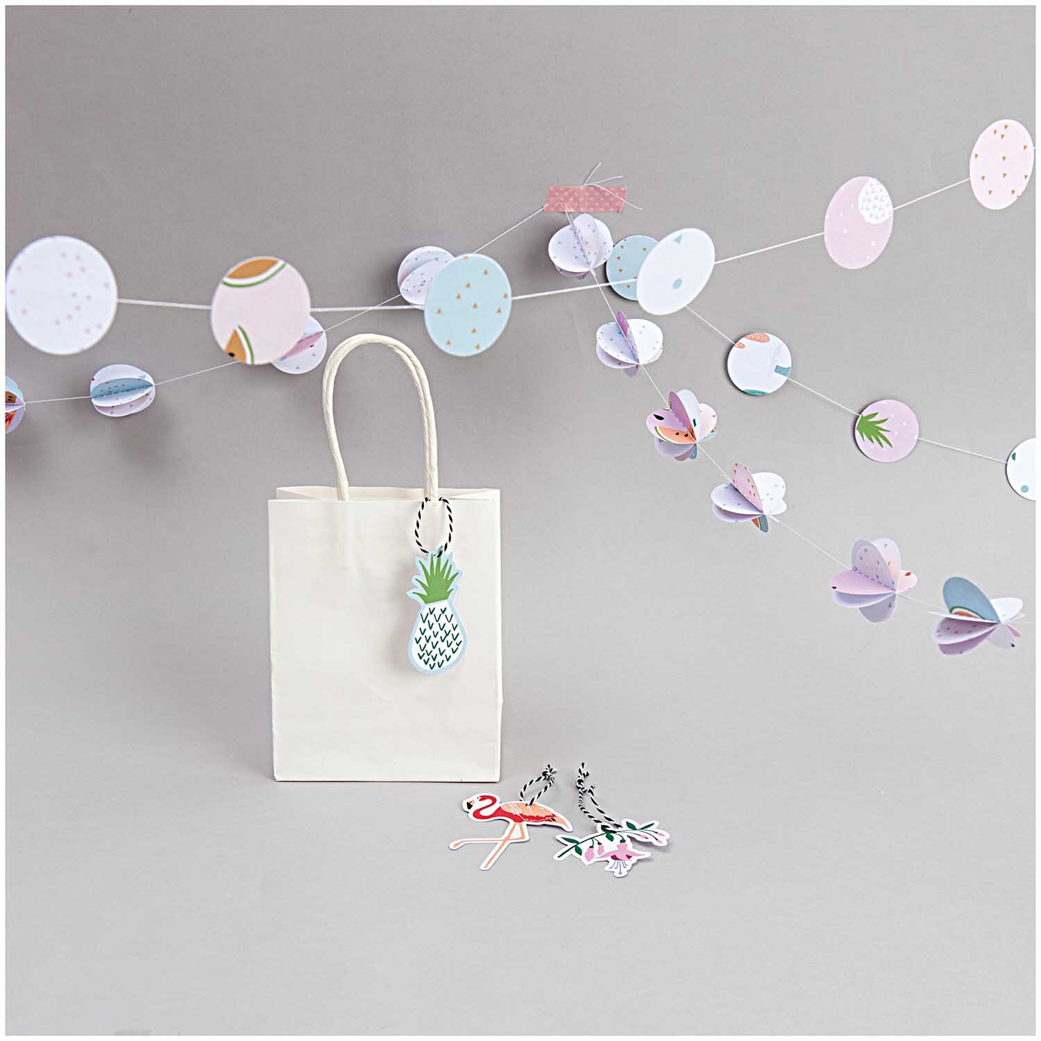 Papiertüte weiß von Rico Design vor grauem Hintergrund mit bunter Girlande und Dekoration.