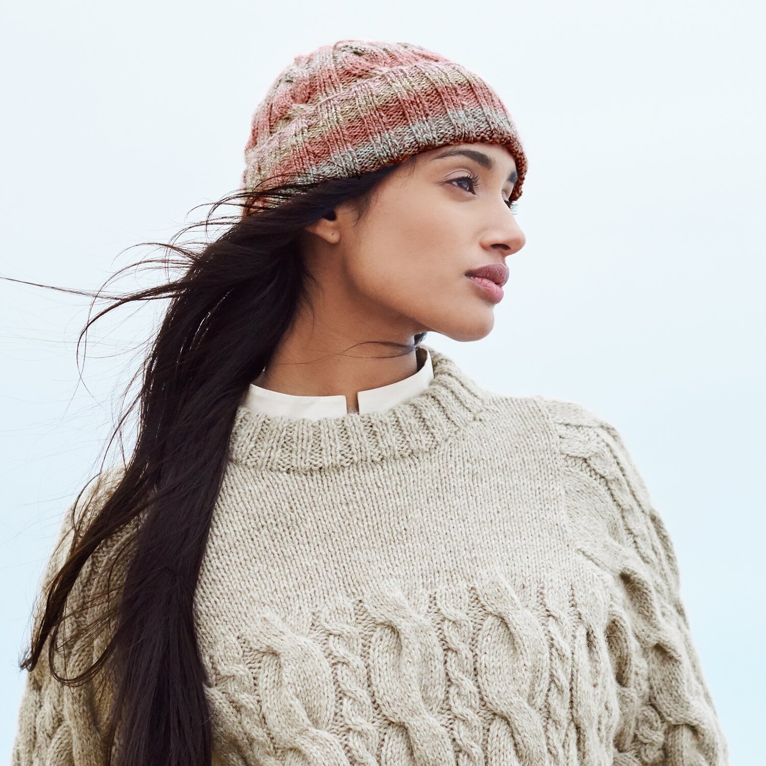 Strickset Mütze Modell 06 aus Lovewool No. 21 von Rico Design Strickset Mütze Modell 06: Frau mit beiger Zopfpullover und bunter Mütze blickt in die Ferne.