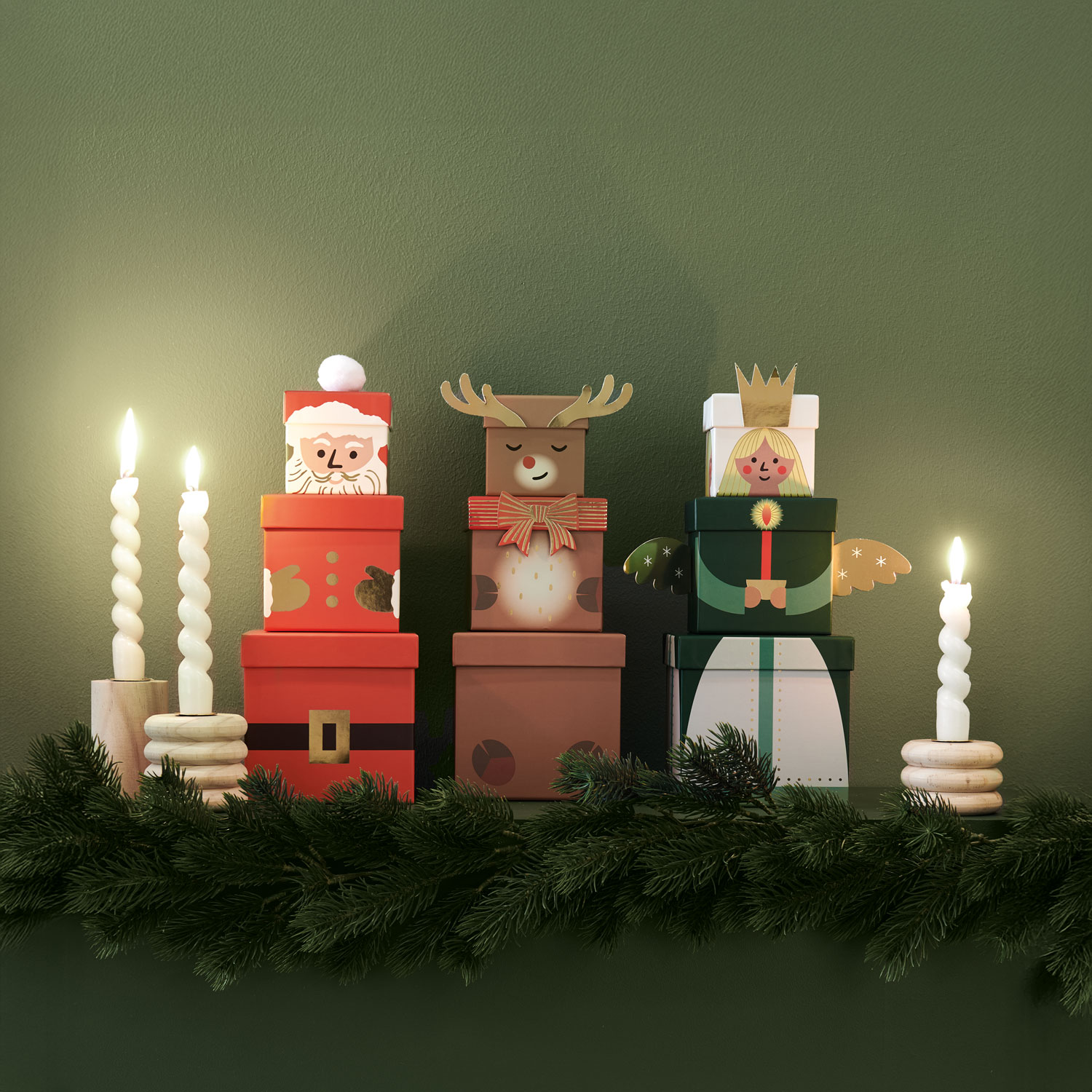 Jeweils drei gestapelte Geschenkschachteln bilden einen Weihnachtsmann, ein Rentier und einen Engel, daneben Stabkerzen. Fotosetting grüner Hintergrund.