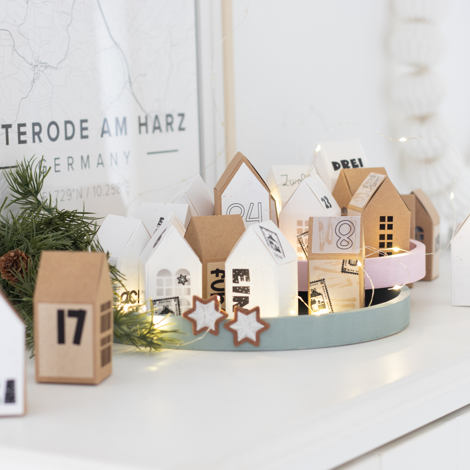 Inspiration Adventskalender Weihnachtsdorf @machsschoen Inspiration Adventskalender Weihnachtsdorf: Dekoratives Dorf aus Papphäusern in Weiß und Braun auf einem Tablett, umgeben von Lichtern.