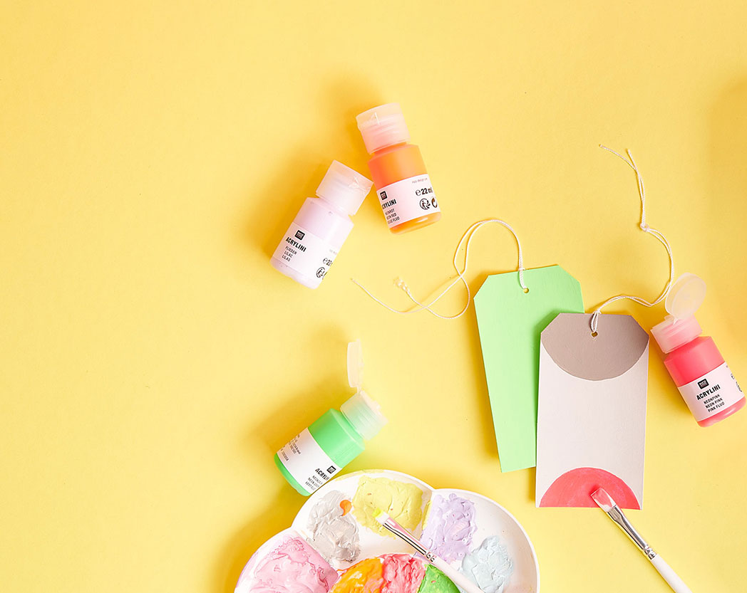 Kleine Acrylfarben Tuben mit einer Malpalette, Pinsel und gestalteten Geschenkanhängern. Flatlay von Produkten.