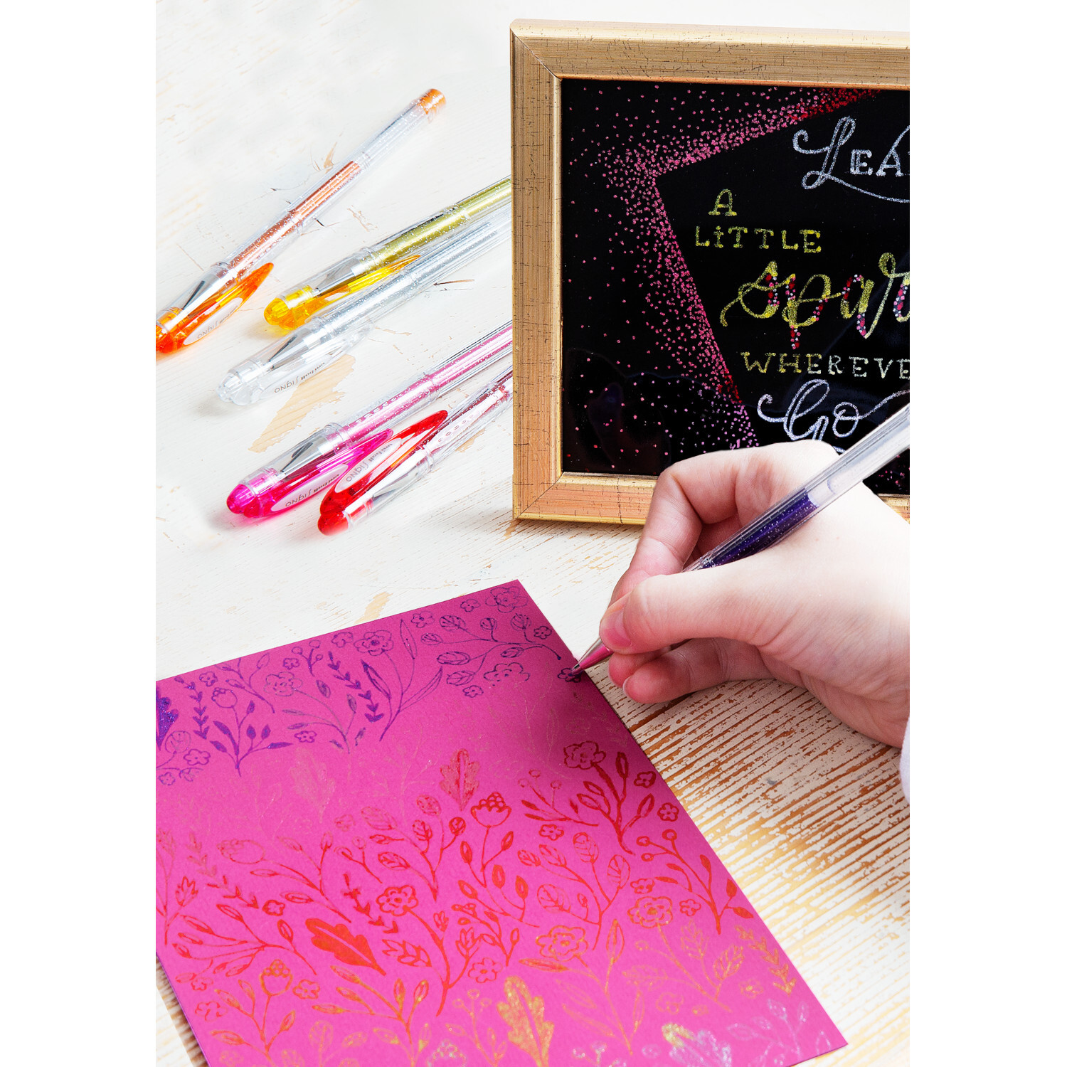 Uni-Ball Signo UM-120AC Gelroller 0,45mm Farbe Orange von Faber-Castell Uni-Ball Signo UM-120AC Gelroller schreibt auf pinkem Papier mit floralen Mustern, umgeben von bunten Stiften und einem dekorativen Rahmen.