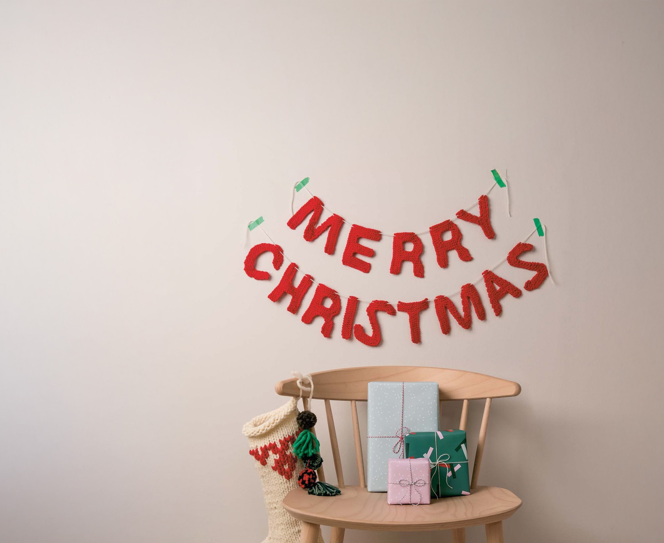 Auf einem Holzstuhl liegen drei verpackte Geschenke und eine Weihnachtssocke, darüber hängt eine rote Girlande mit weihnachtlichem Spruch.