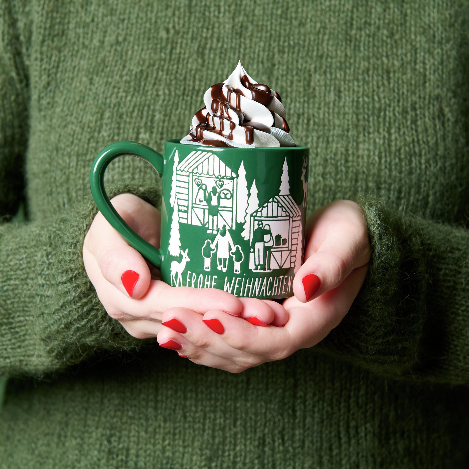 Porzellan Tasse Weihnachtsmarkt mit Sahne und Schokosoße, gehalten von Händen in grünem Pullover.