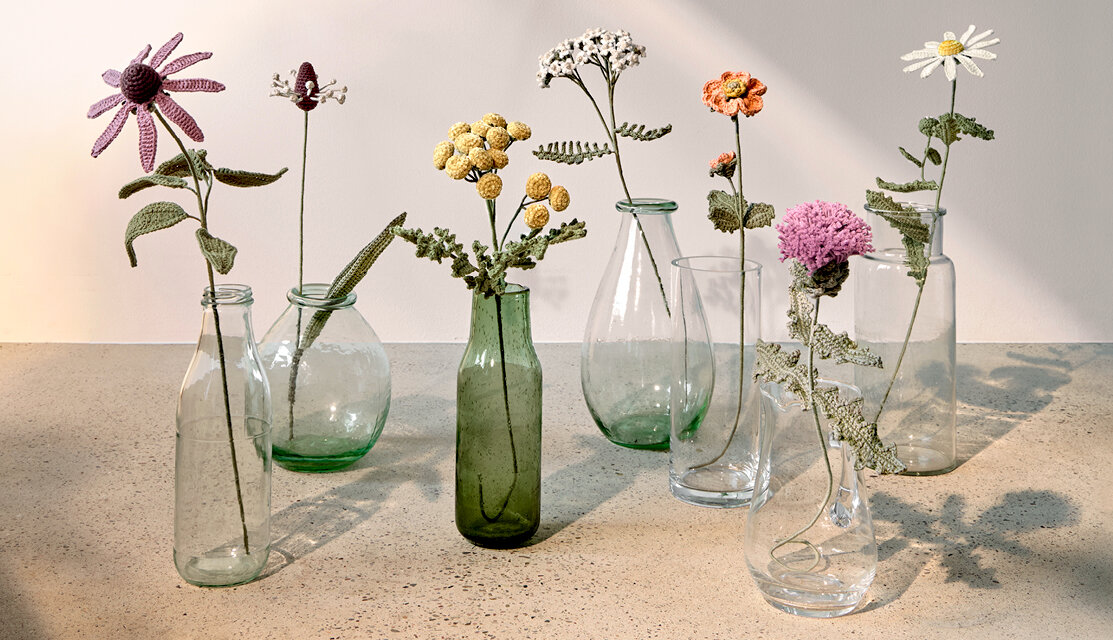 Material Ricorumi In the Meadow Ackerdistel, gehäkelte Wiesenblumen in transparenten Glasvasen, arrangiert auf hellem Steintisch in natürlichem Licht